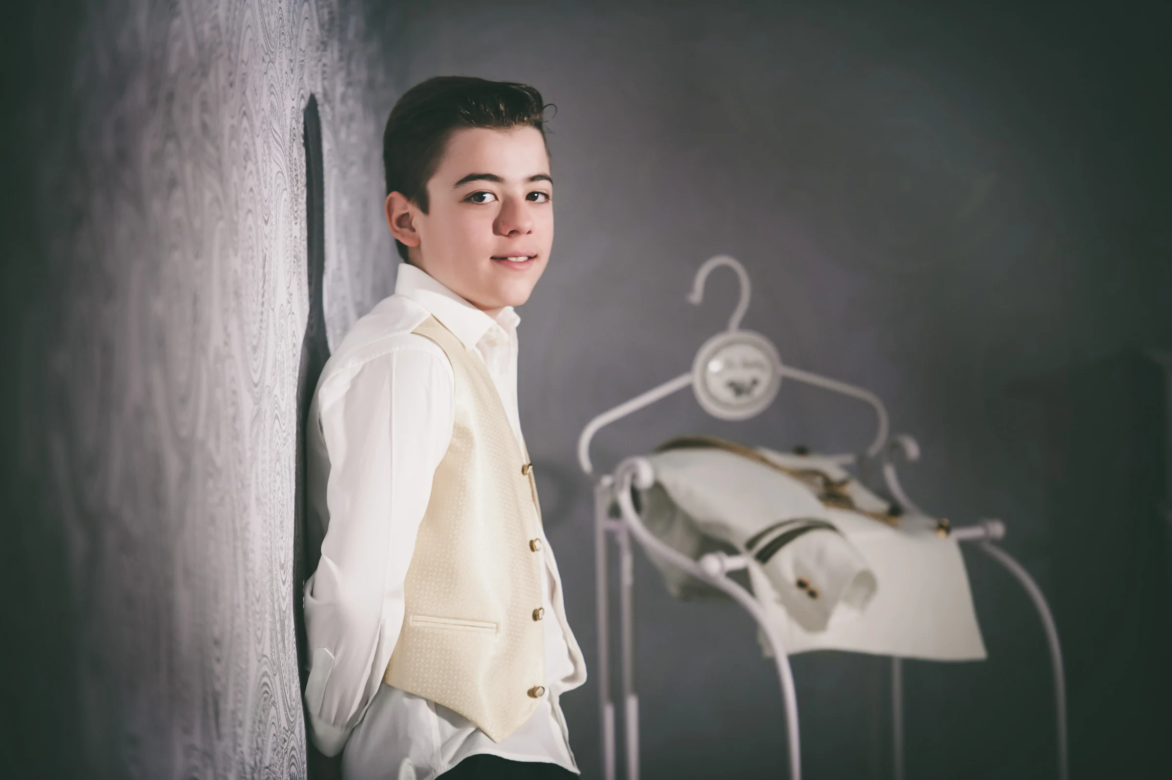Niño con camisa blanca y chaleco beige posa junto a su traje de Primera Comunión colgado en el estudio.