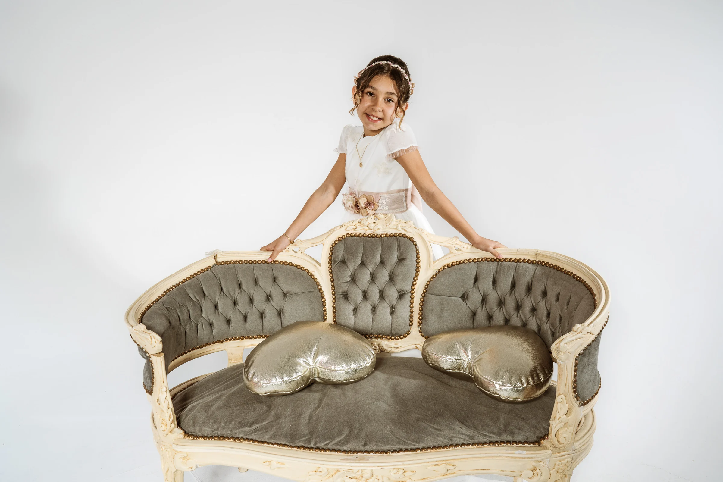 Niña con vestido de Primera Comunión posa detrás de un sofá clásico de capitoné en un estudio de fondo blanco.