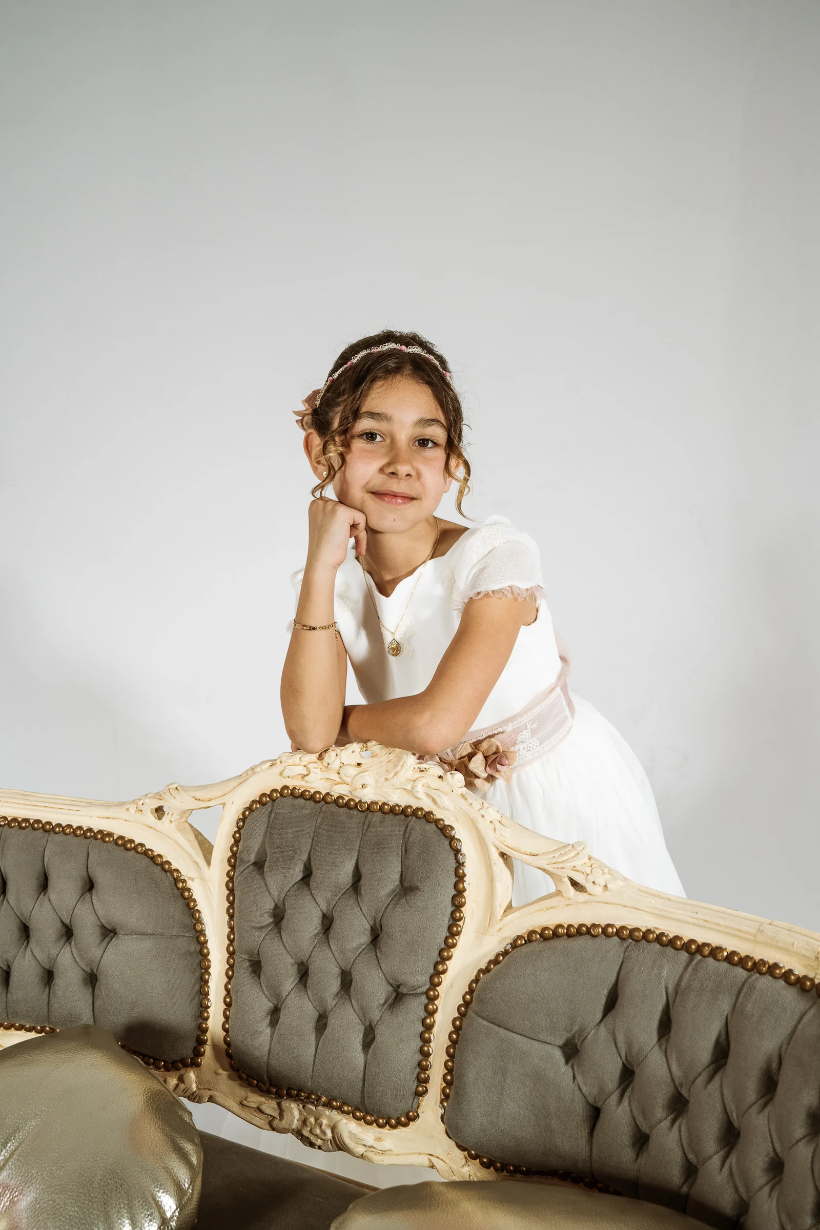 Niña con vestido de Primera Comunión se apoya sobre el respaldo de un sofá clásico en un retrato de estudio.