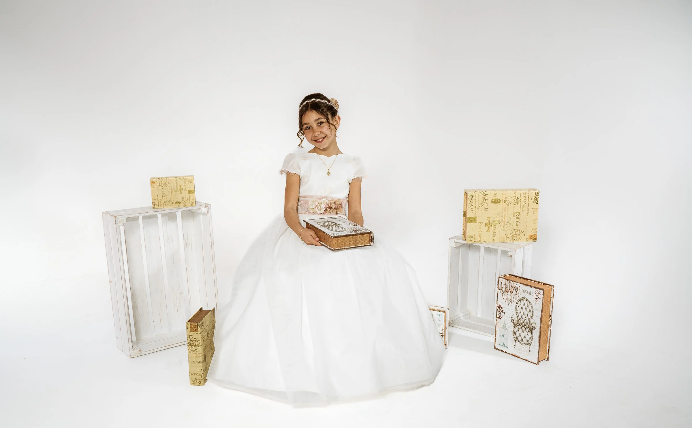 Niña con vestido de Primera Comunión posa sentada entre cajas blancas y libros decorativos sobre fondo claro.