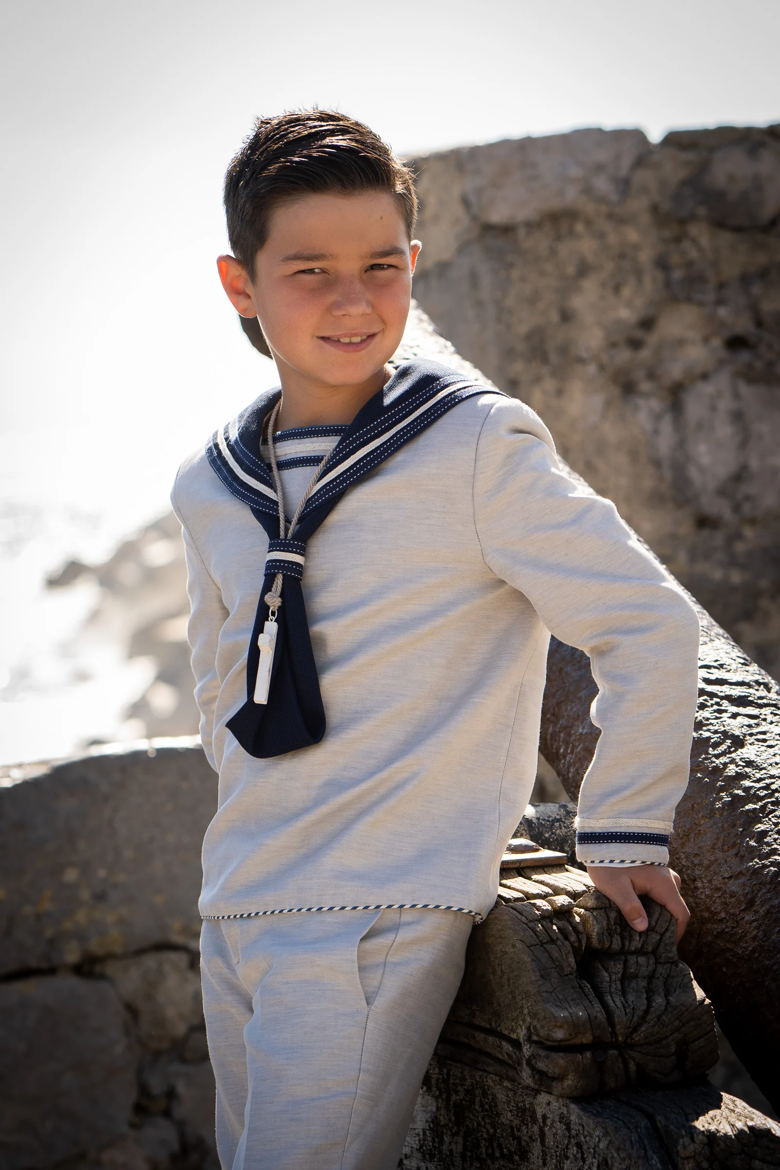 Niño con traje de Primera Comunión de estilo marinero posa junto a un muro de piedra con el mar al fondo.