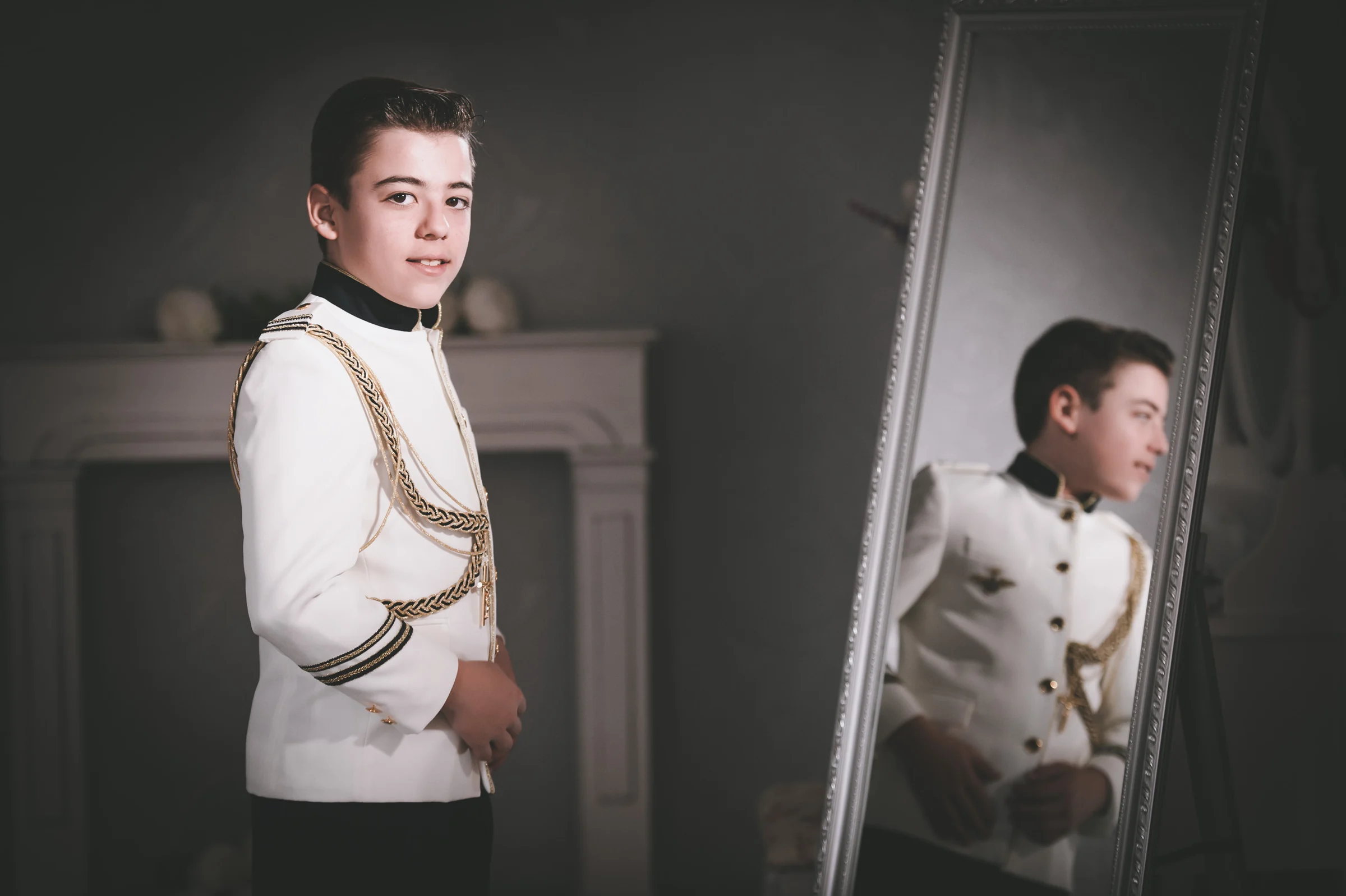 Niño con traje blanco de Primera Comunión posa junto a un gran espejo que refleja su figura en el estudio.