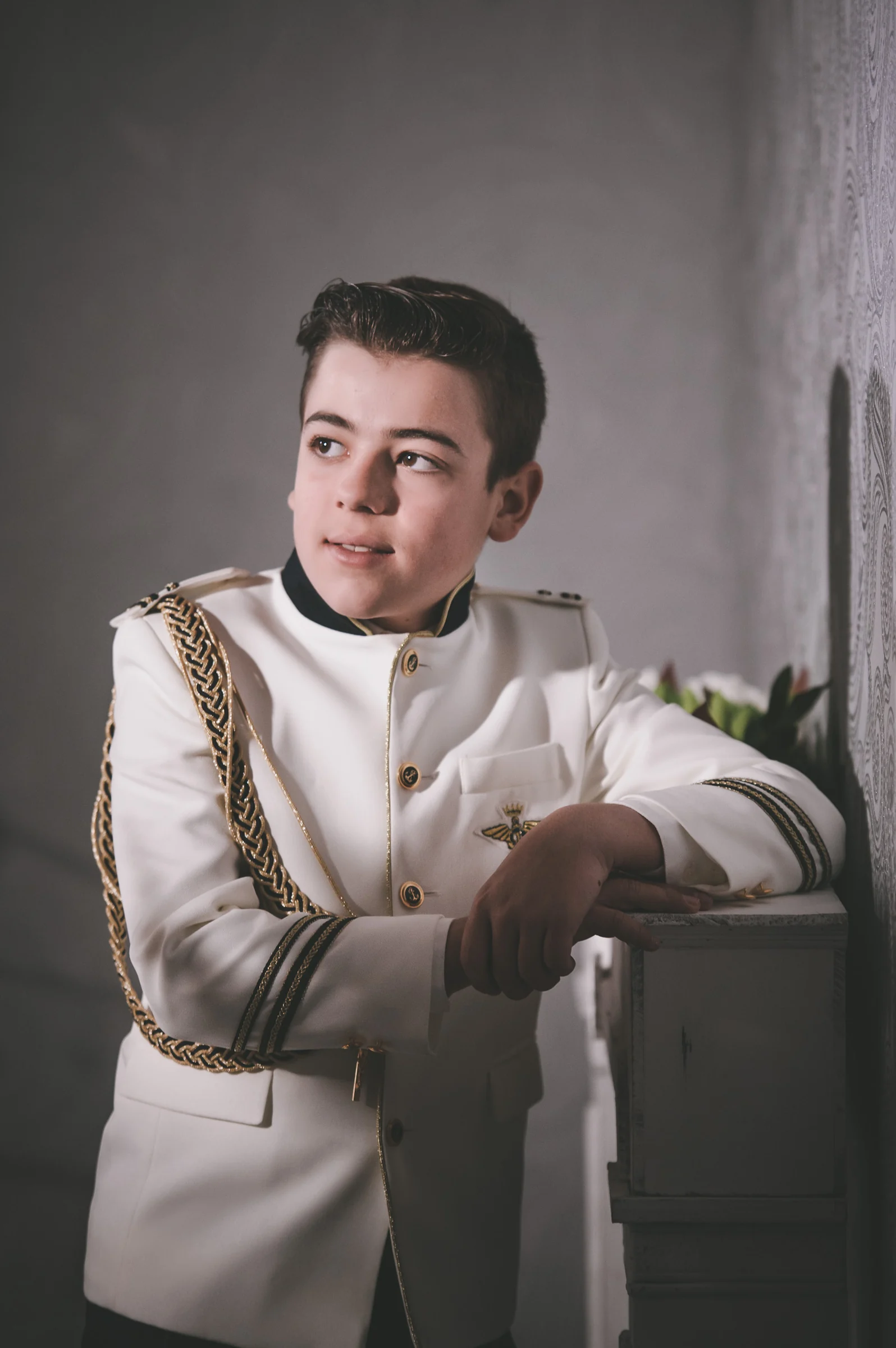 Niño con traje blanco de Primera Comunión se apoya sobre un mueble claro en un retrato de luz suave.