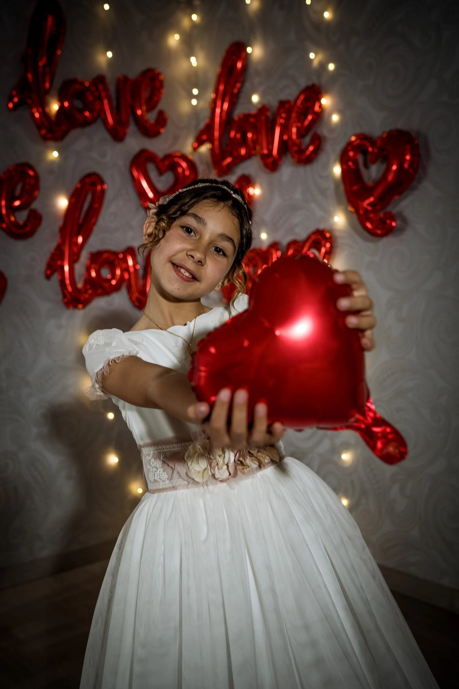 Niña con vestido de Primera Comunión sostiene un globo rojo con forma de corazón ante luces y decoración de corazones.