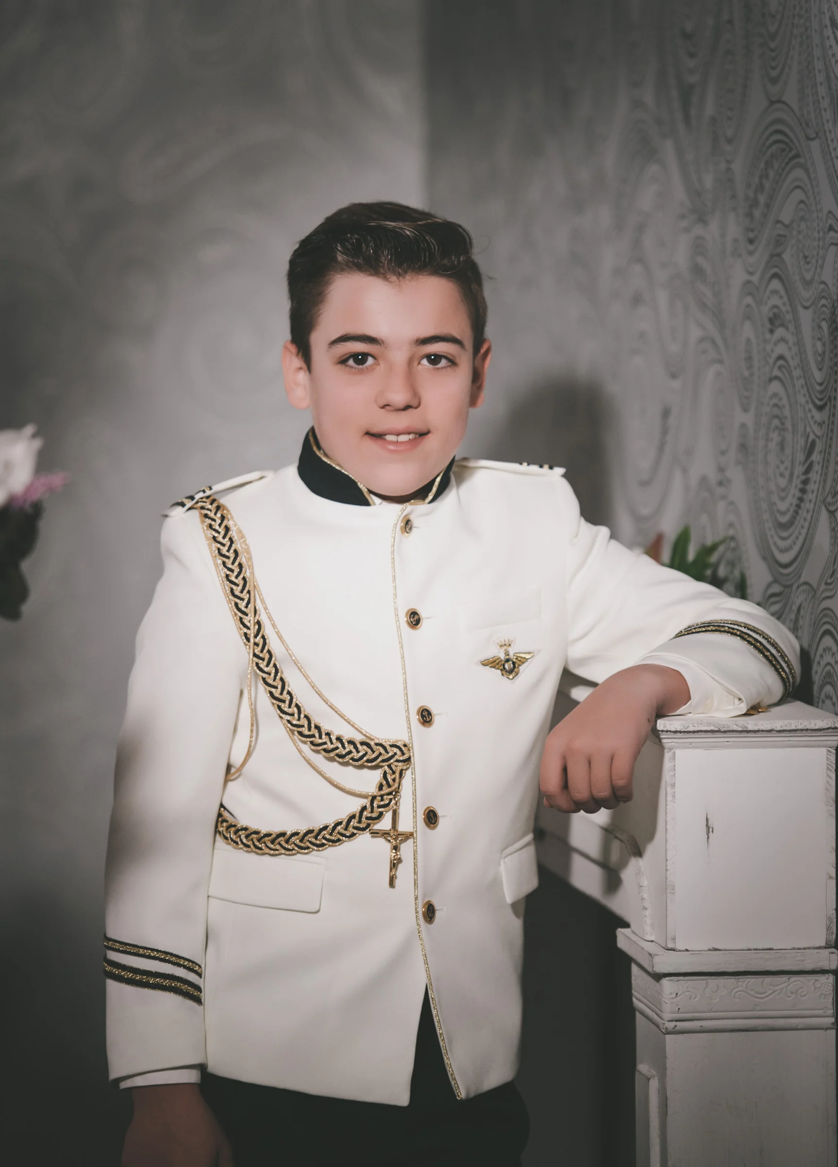 Niño con traje blanco de Primera Comunión y detalles dorados posa apoyado sobre una columna en estudio.