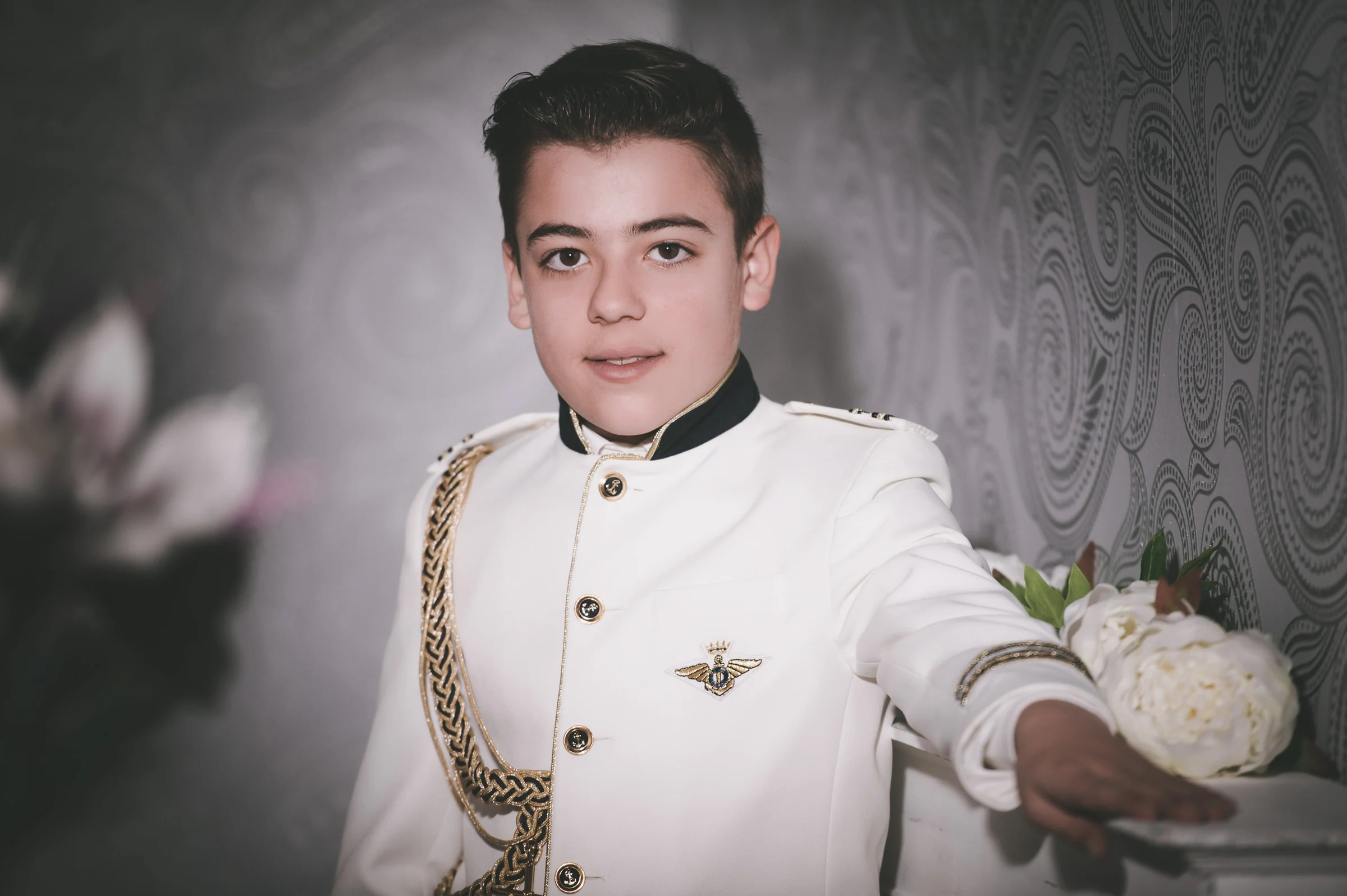 Retrato cercano de niño con traje blanco de Primera Comunión y cordones dorados junto a flores claras.