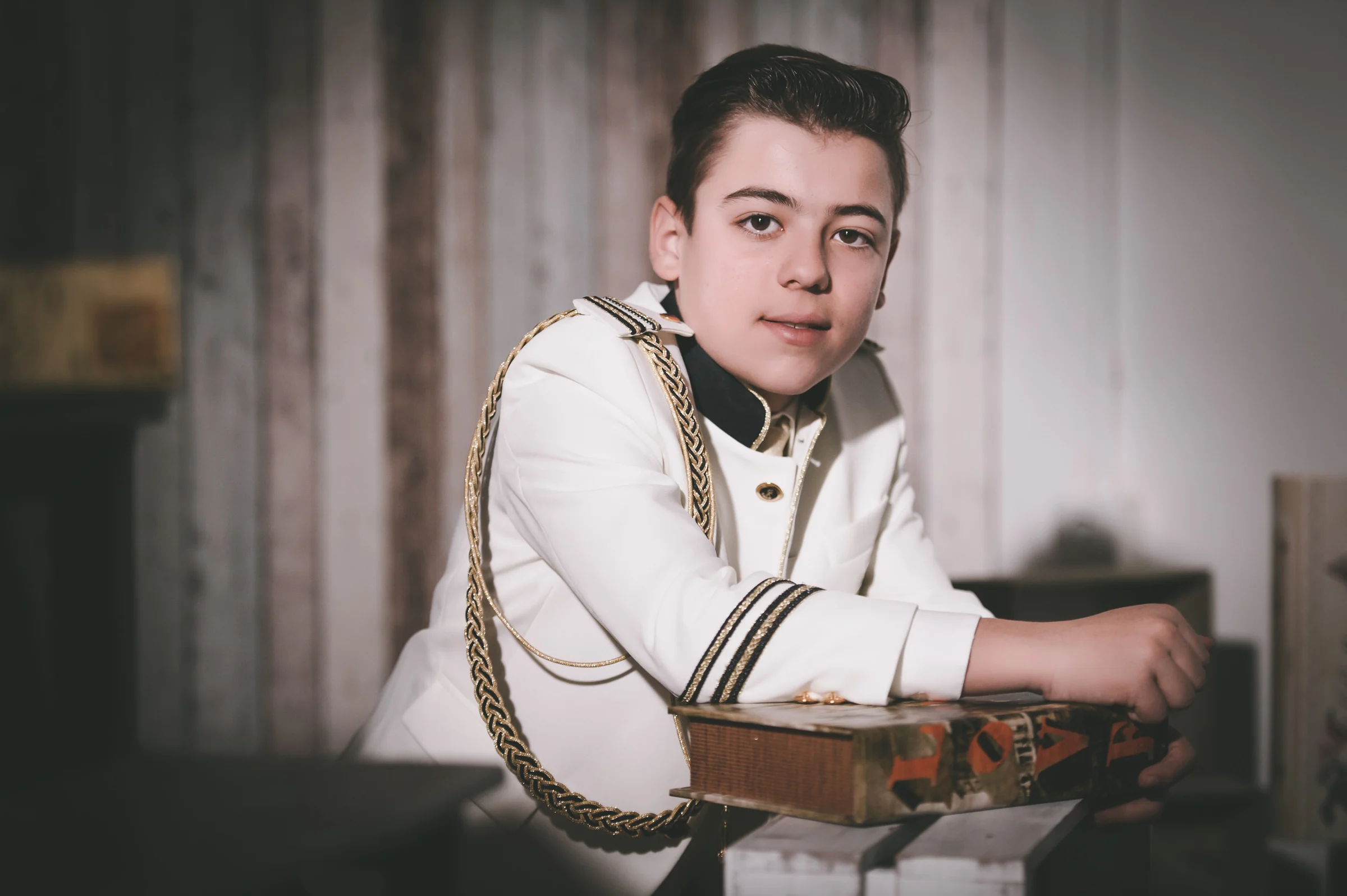 Niño con traje blanco de Primera Comunión se apoya sobre una caja decorativa en un estudio de ambiente oscuro.