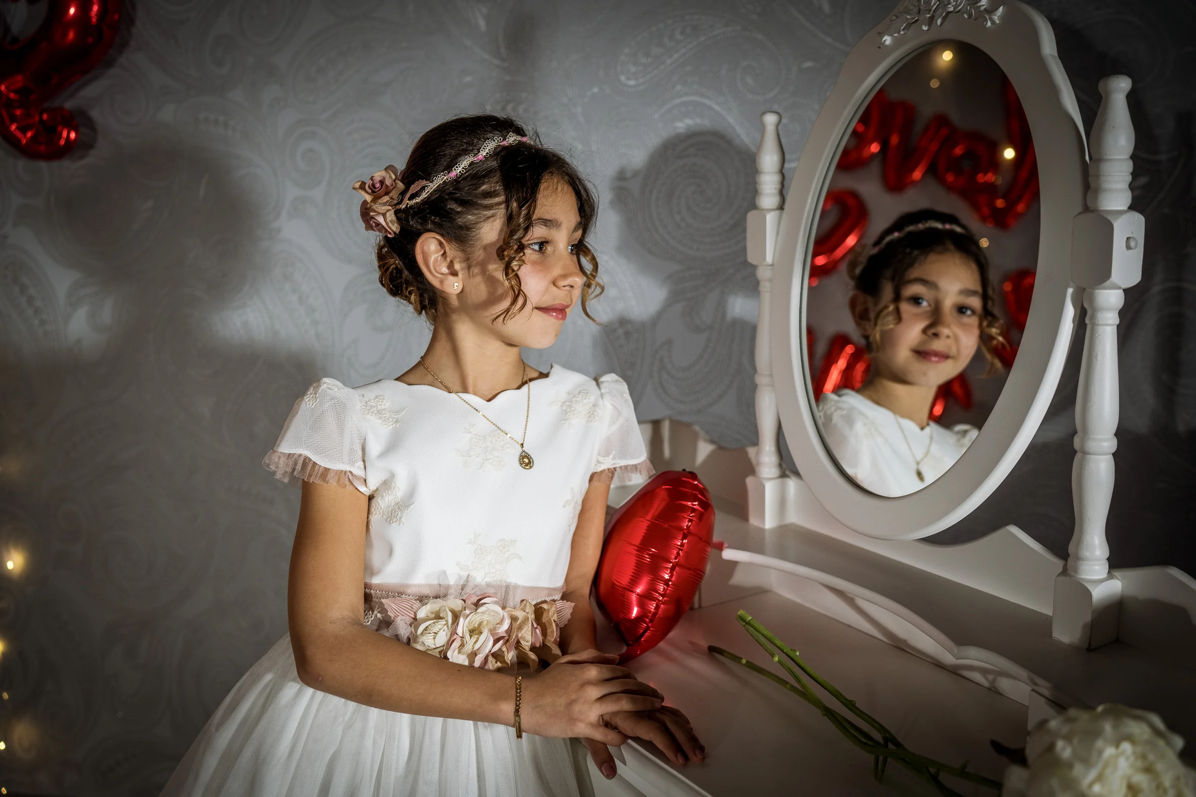 Niña con vestido de Primera Comunión posa junto a un tocador y un espejo sosteniendo un globo rojo de corazón.