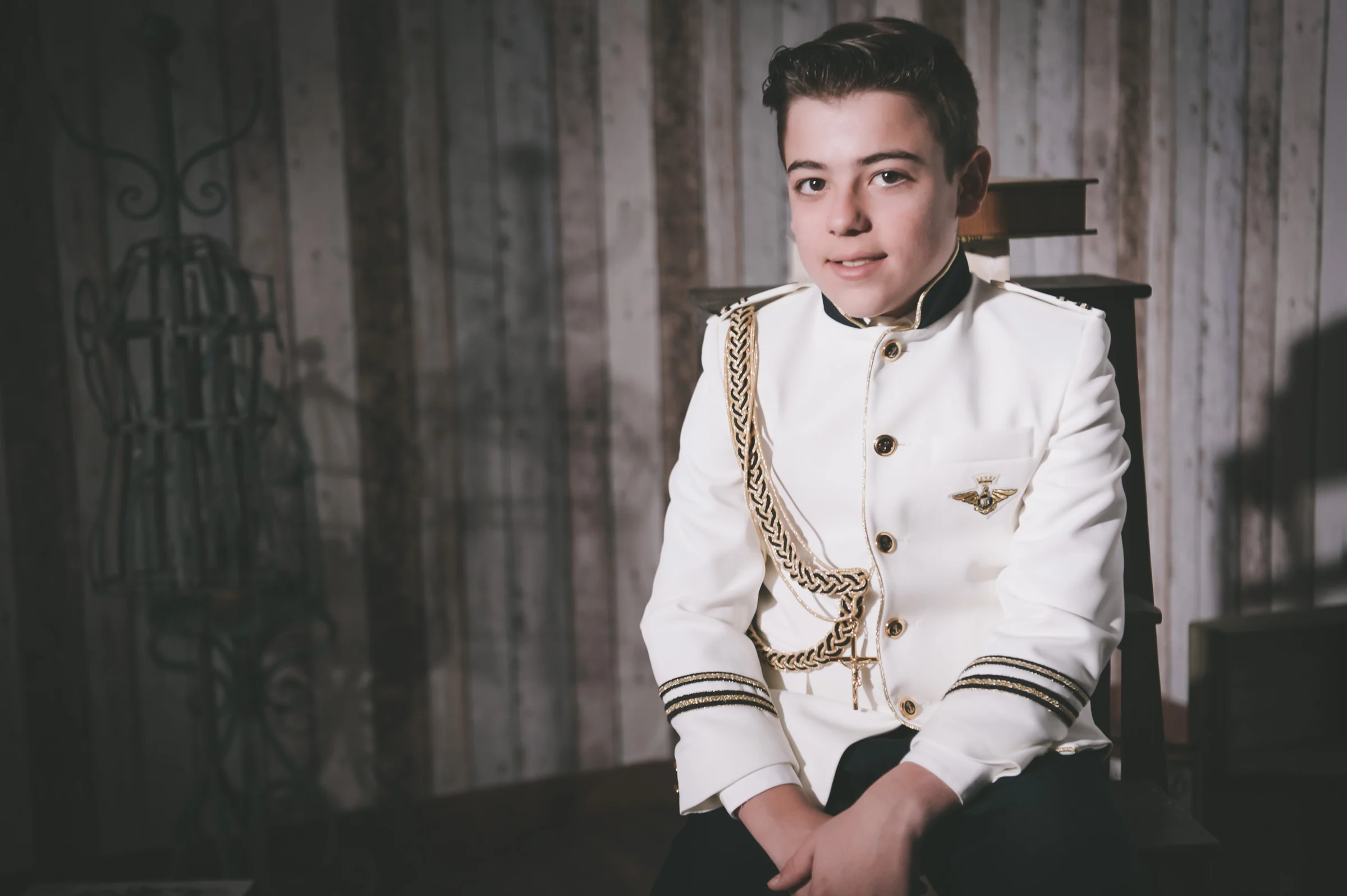 Niño con traje blanco de Primera Comunión posa sentado ante un fondo de madera en una escena de estudio.