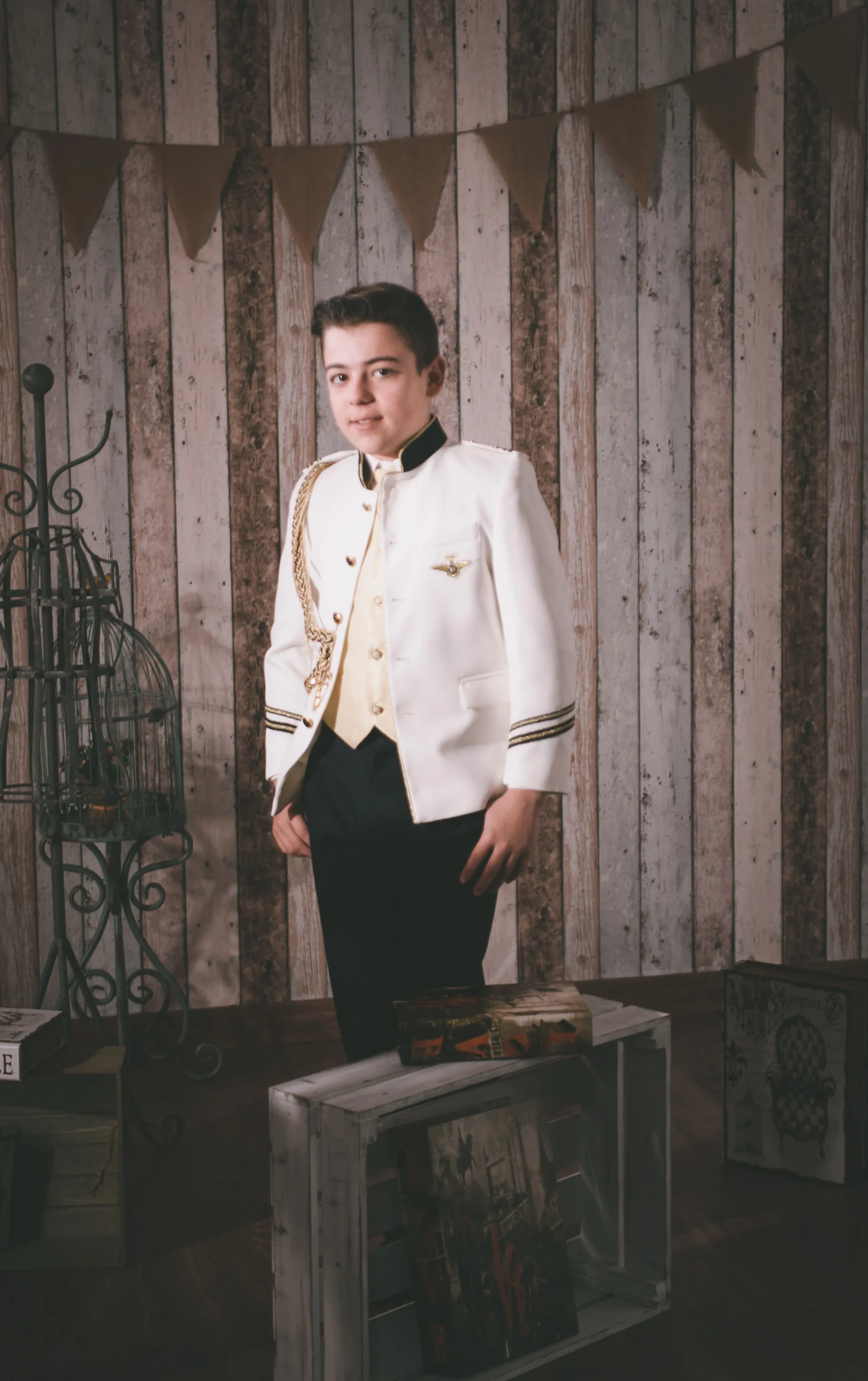 Niño con traje blanco de Primera Comunión posa de cuerpo entero ante un fondo rústico de madera.