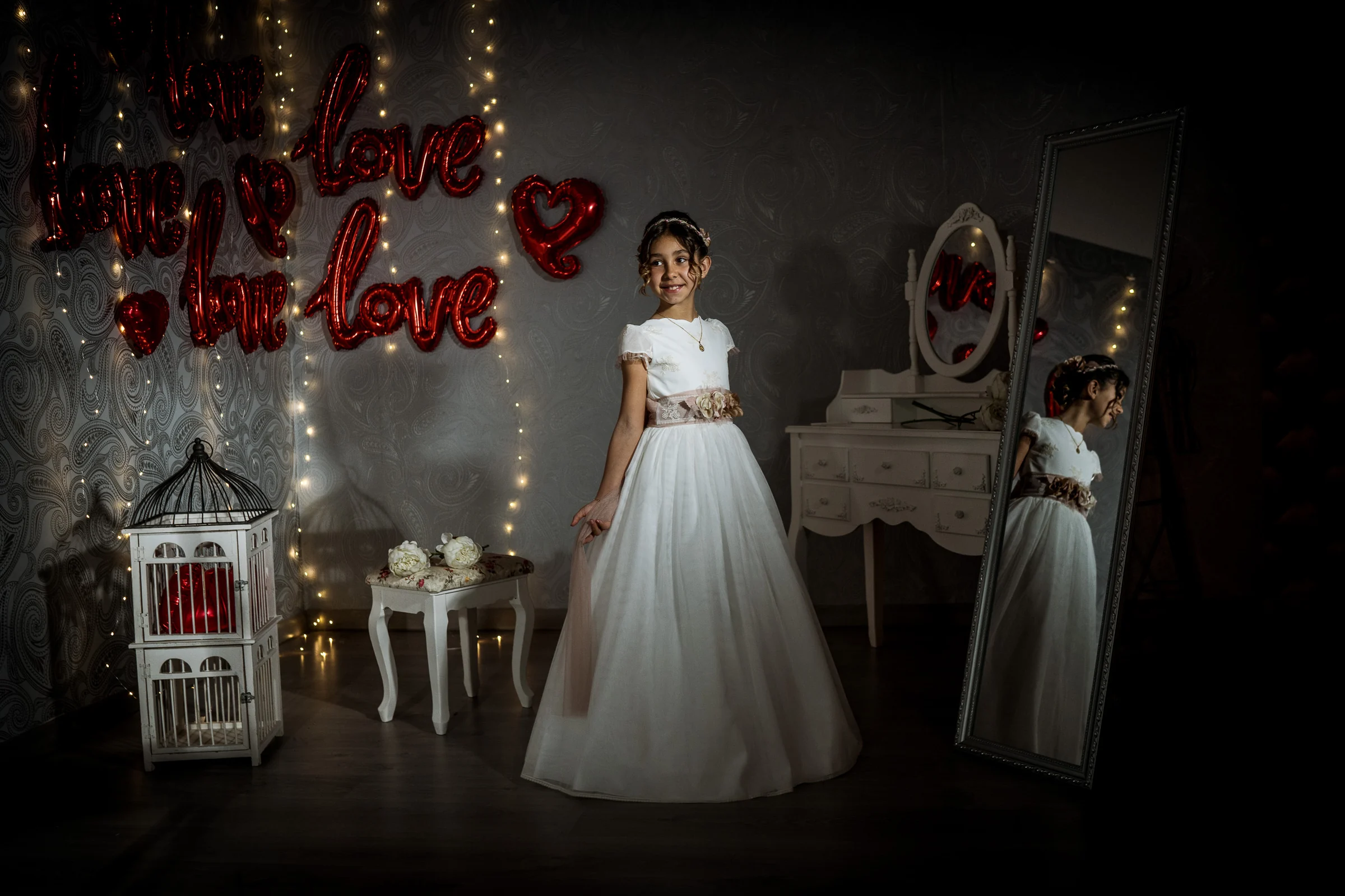 Niña con vestido de Primera Comunión posa de cuerpo entero en un escenario oscuro con luces, corazones y espejo.
