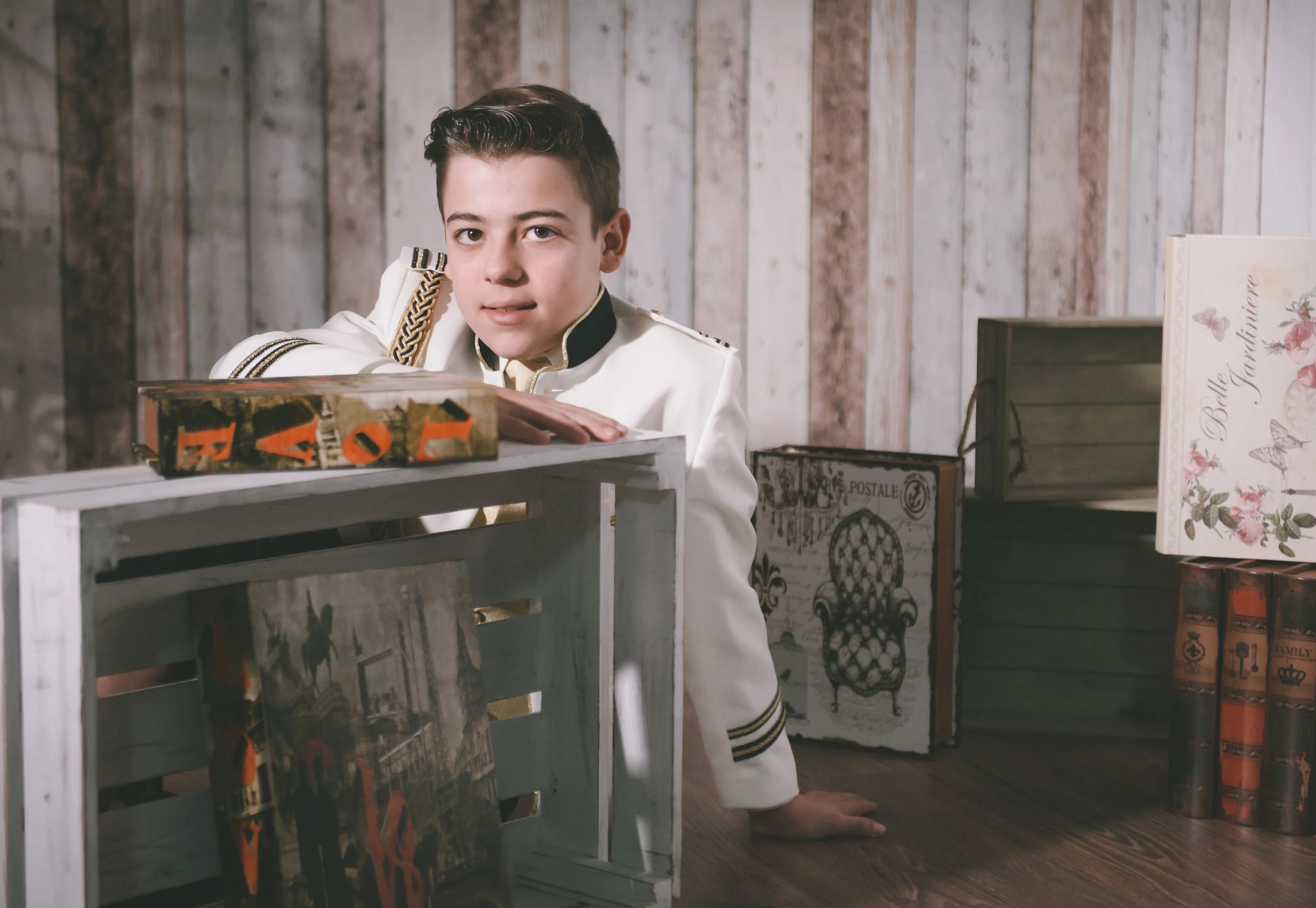 Niño con traje blanco de Primera Comunión posa entre cajas y libros decorativos en un estudio de estilo vintage.