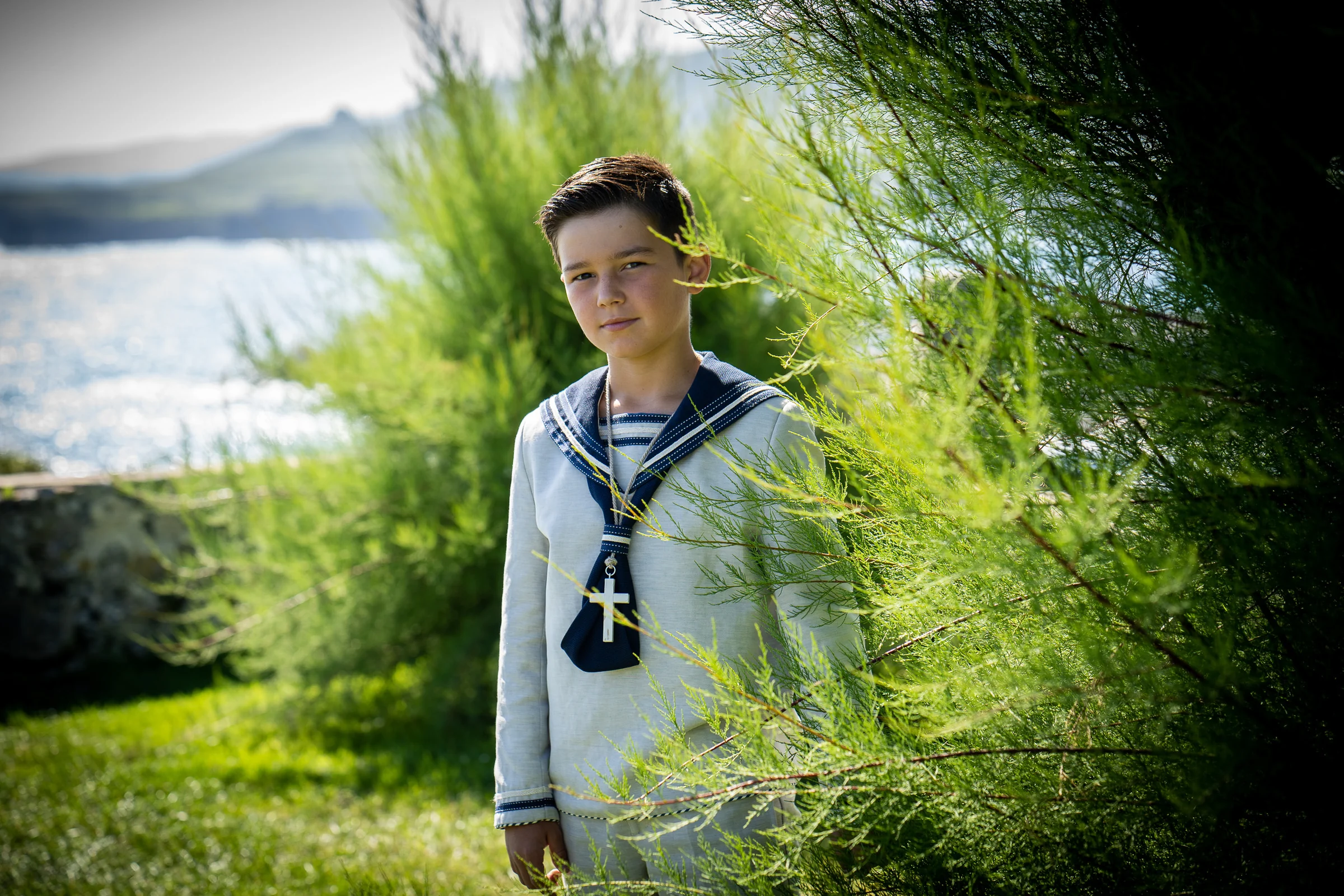 Niño con traje de Primera Comunión de estilo marinero posa entre vegetación verde con el mar al fondo.