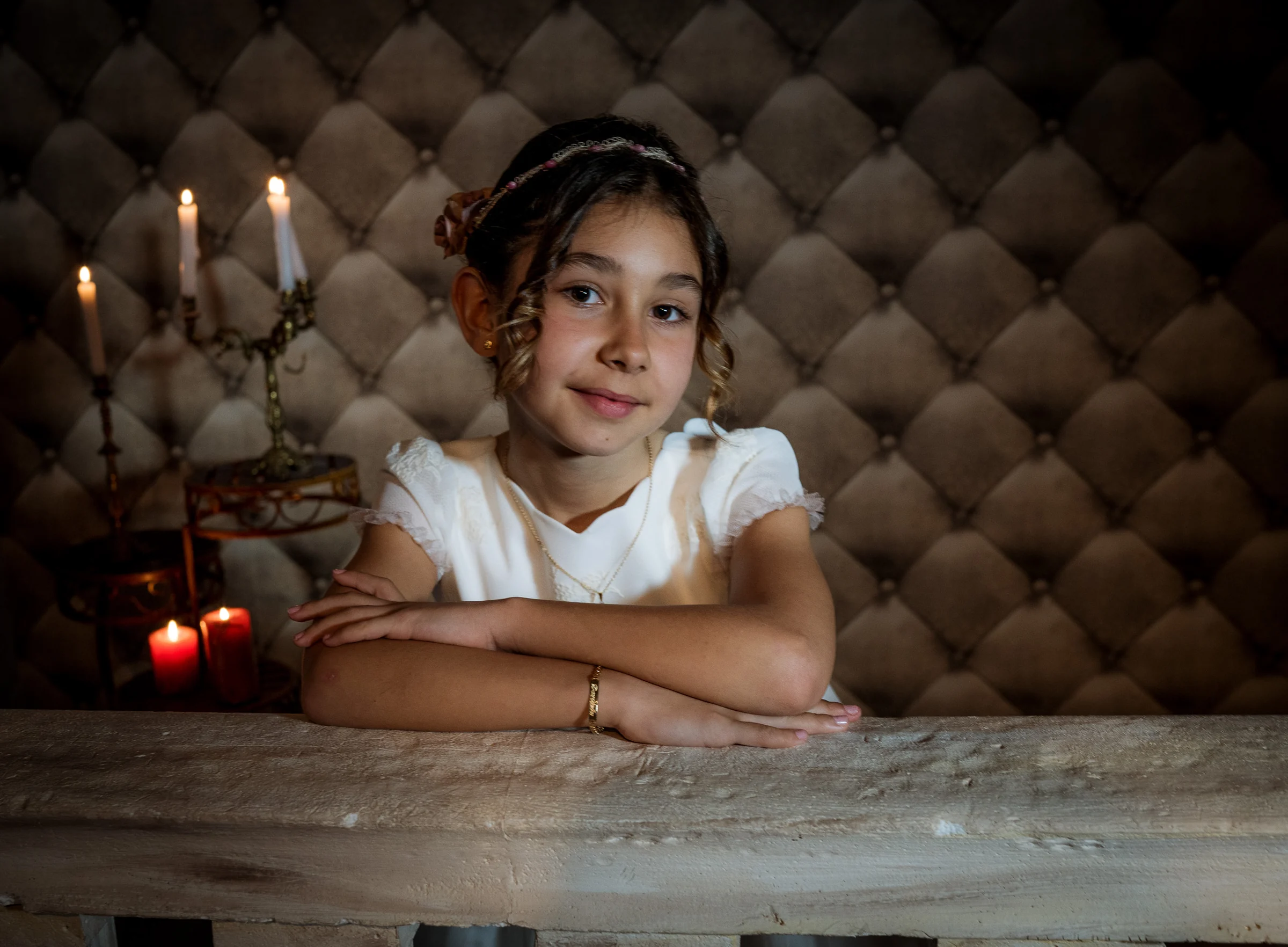 Niña con vestido de Primera Comunión apoya los brazos sobre una mesa en un retrato con velas y fondo capitoné.