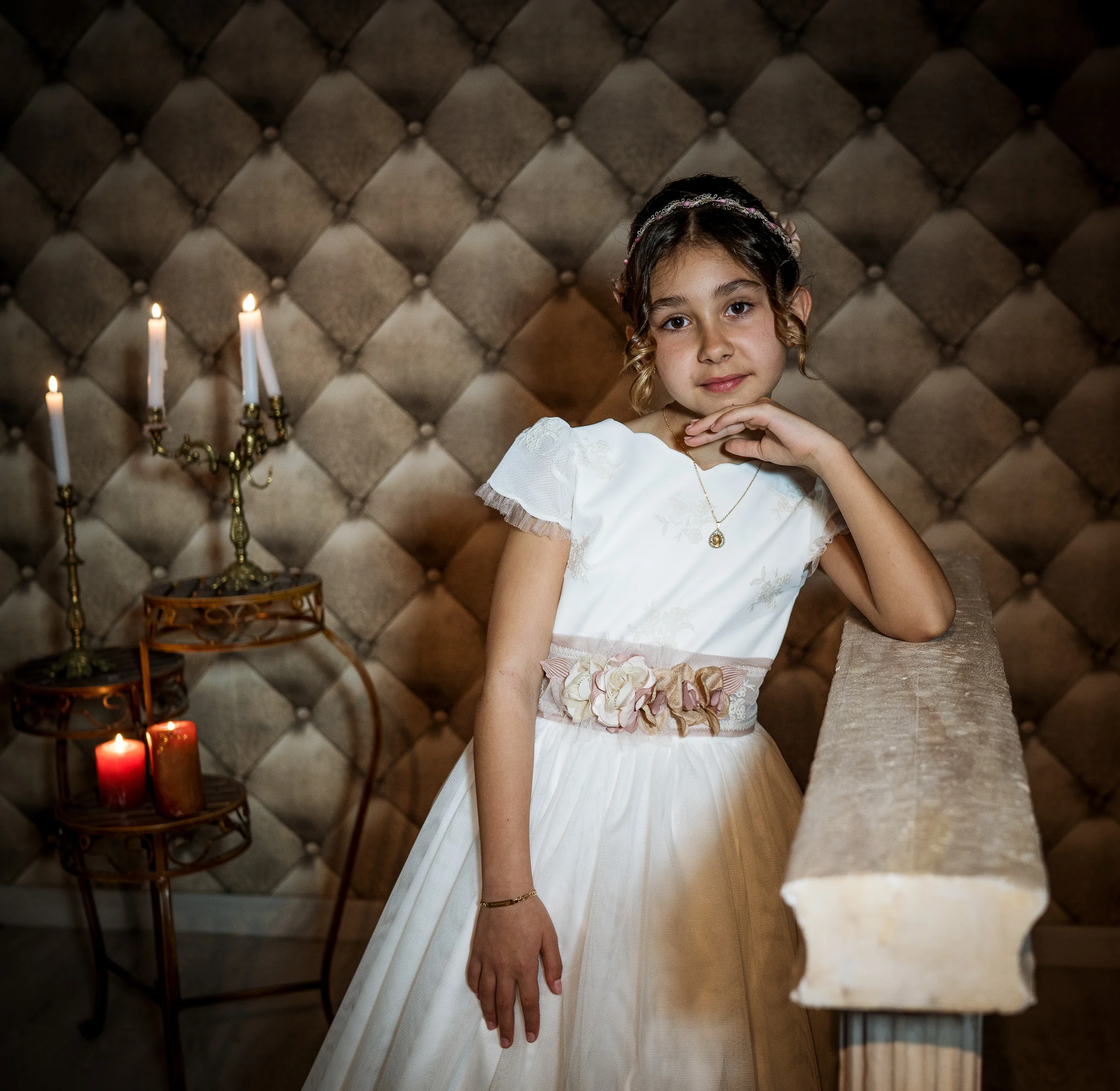 Niña con vestido de Primera Comunión posa junto a una columna decorativa con velas encendidas al fondo.
