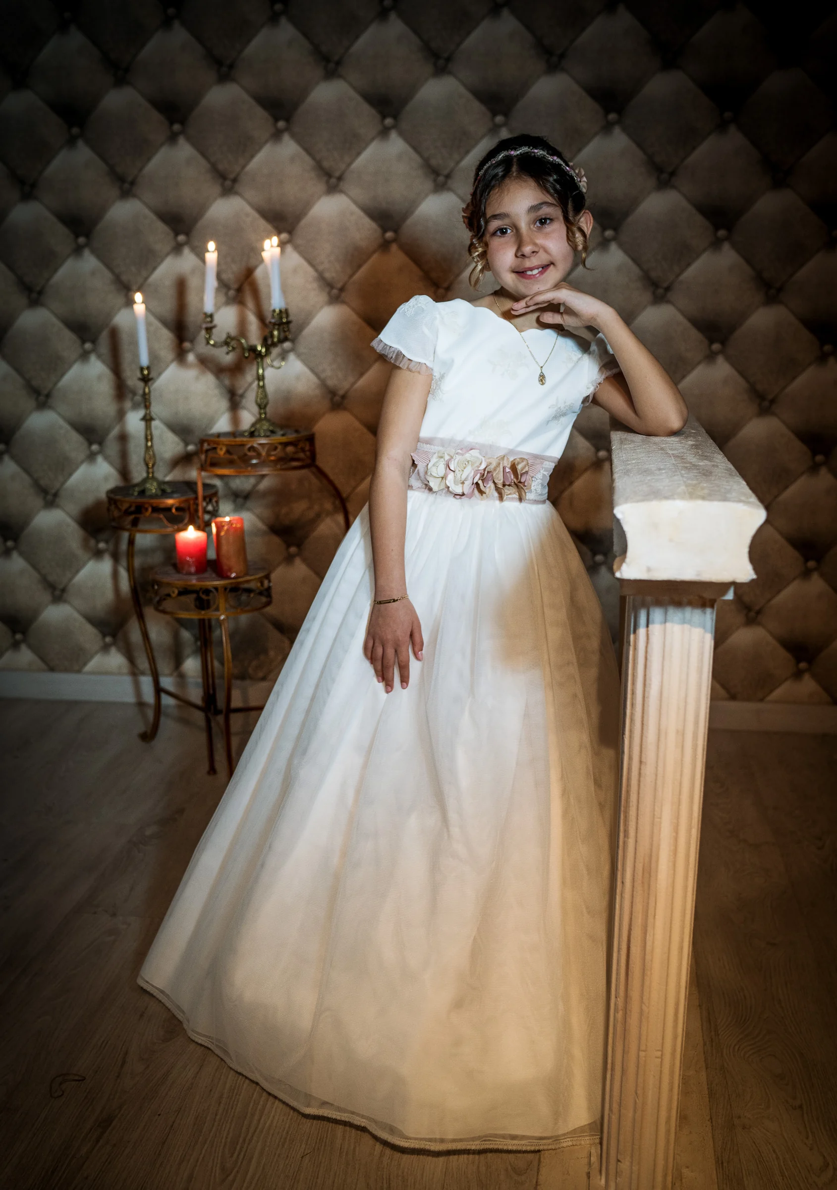 Niña con vestido de Primera Comunión posa de cuerpo entero junto a una columna con un candelabro encendido al fondo.