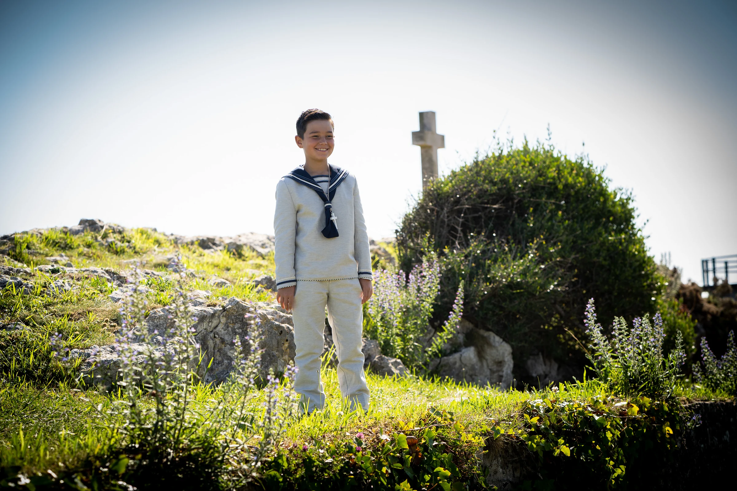 Niño con traje de Primera Comunión de estilo marinero posa al aire libre cerca de una cruz de piedra entre vegetación.