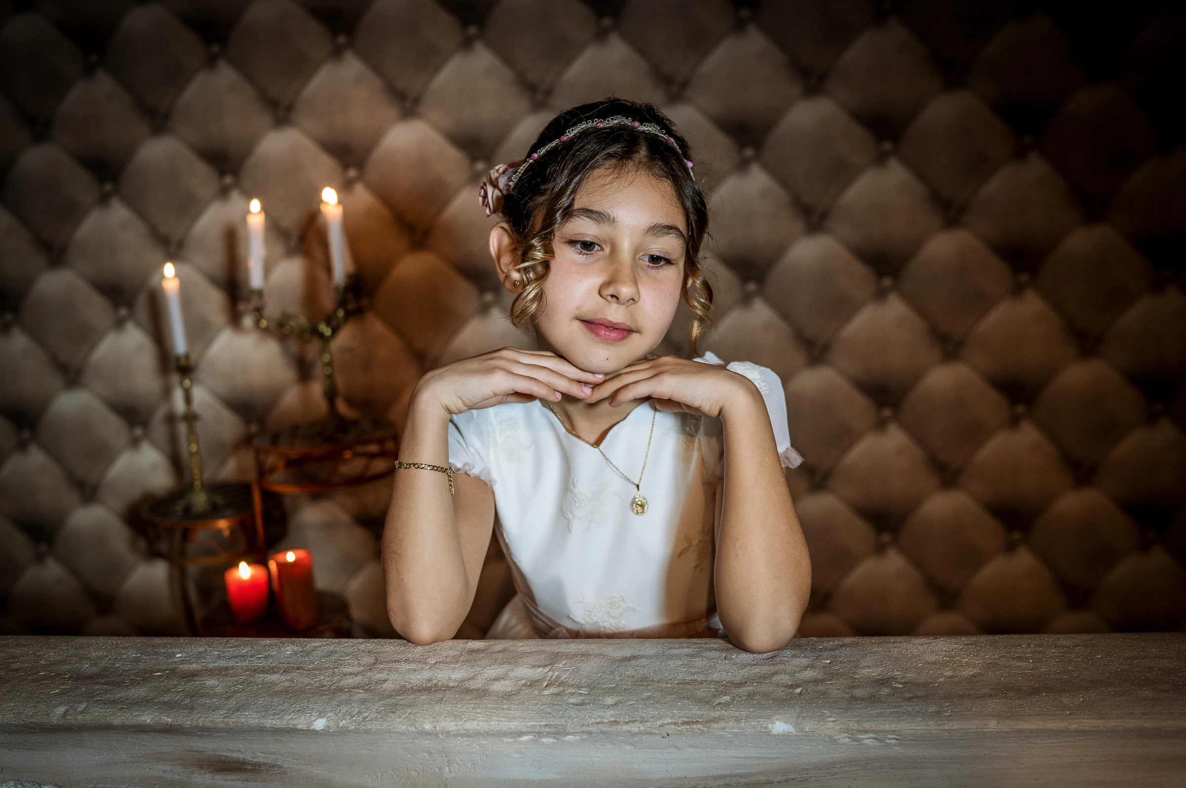 Niña con vestido de Primera Comunión posa con las manos bajo la barbilla sobre una mesa y velas encendidas al fondo.