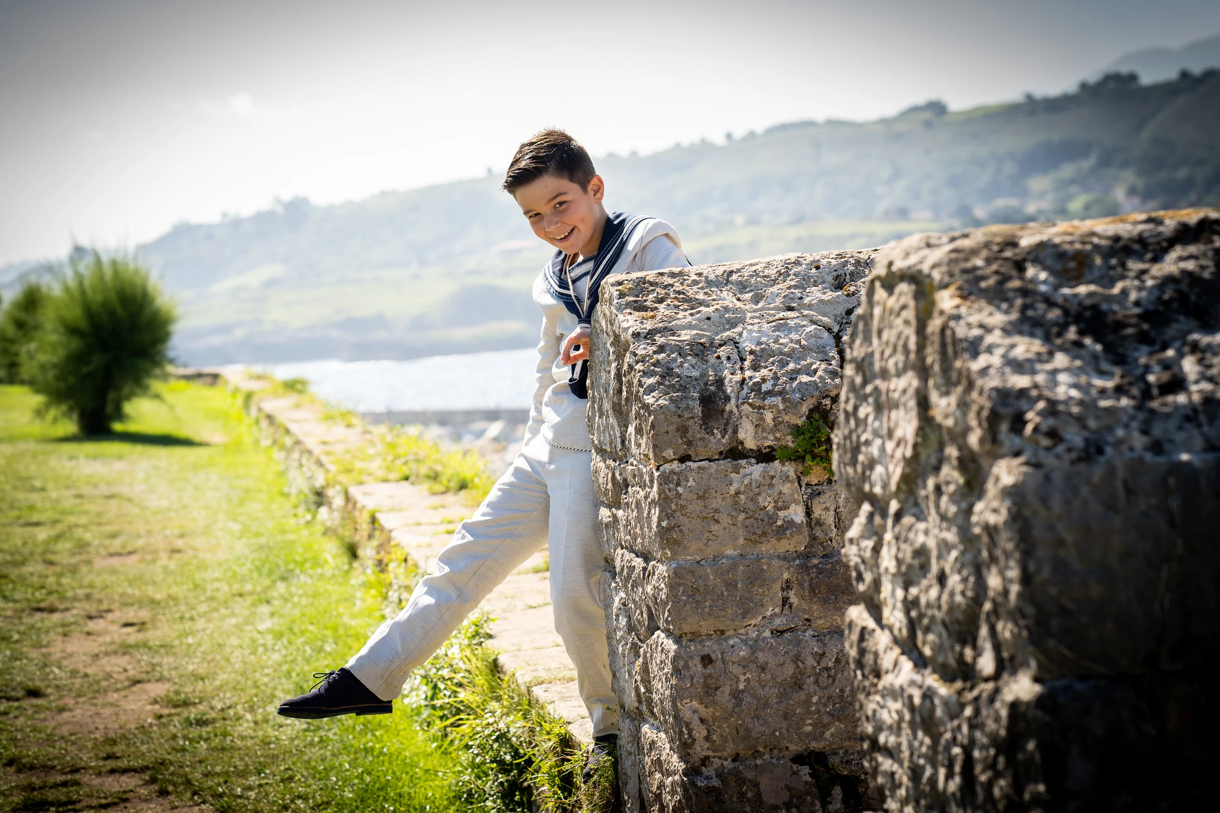 Niño con traje de Primera Comunión juega junto a un muro de piedra en un entorno exterior próximo a la costa.