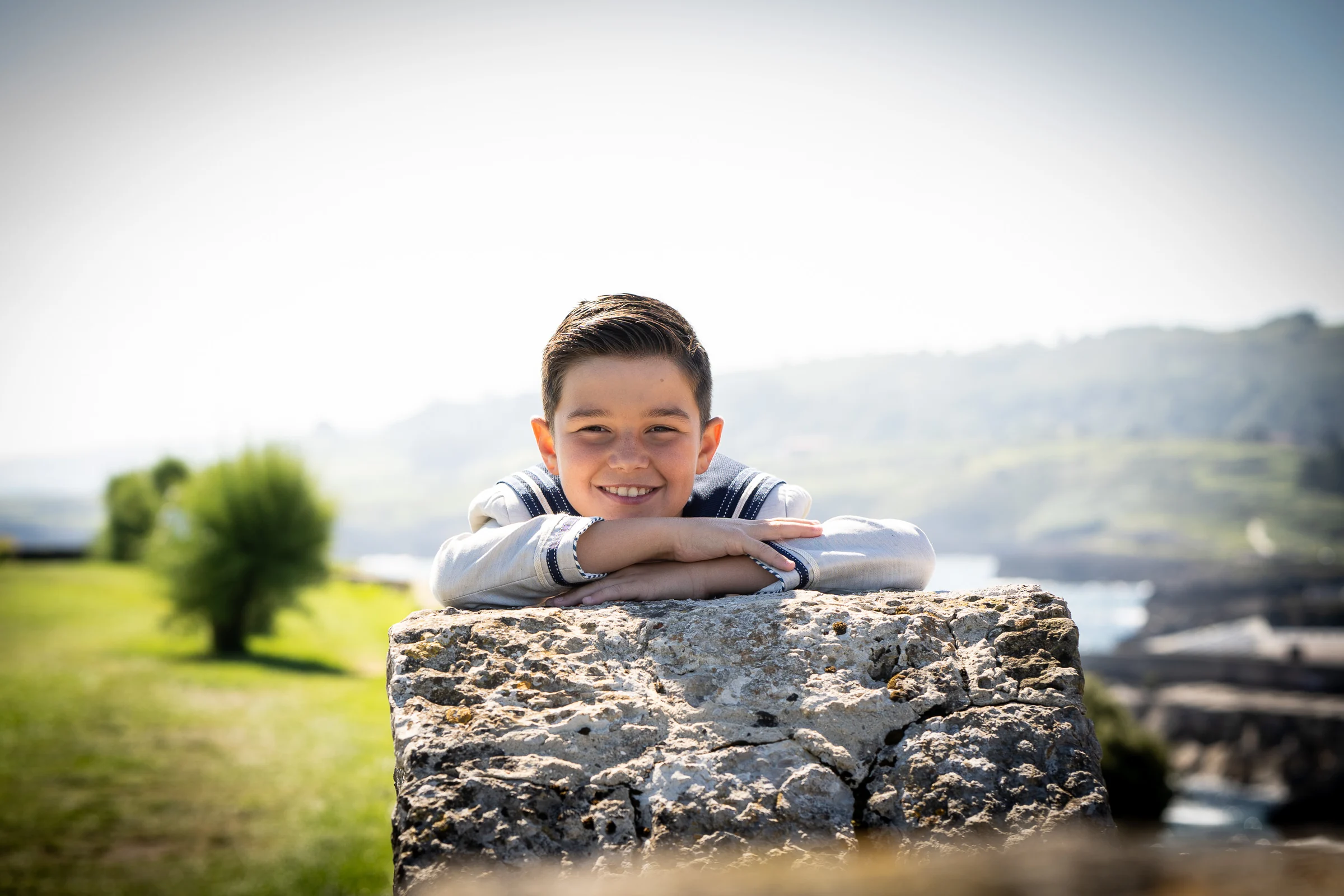 Niño de Primera Comunión sonríe apoyado con los brazos sobre un muro de piedra en un retrato exterior.