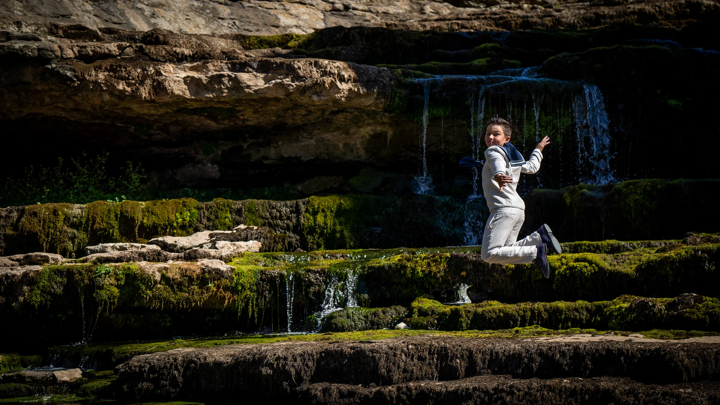 Niño con traje de Primera Comunión salta frente a una pequeña cascada entre rocas cubiertas de musgo.