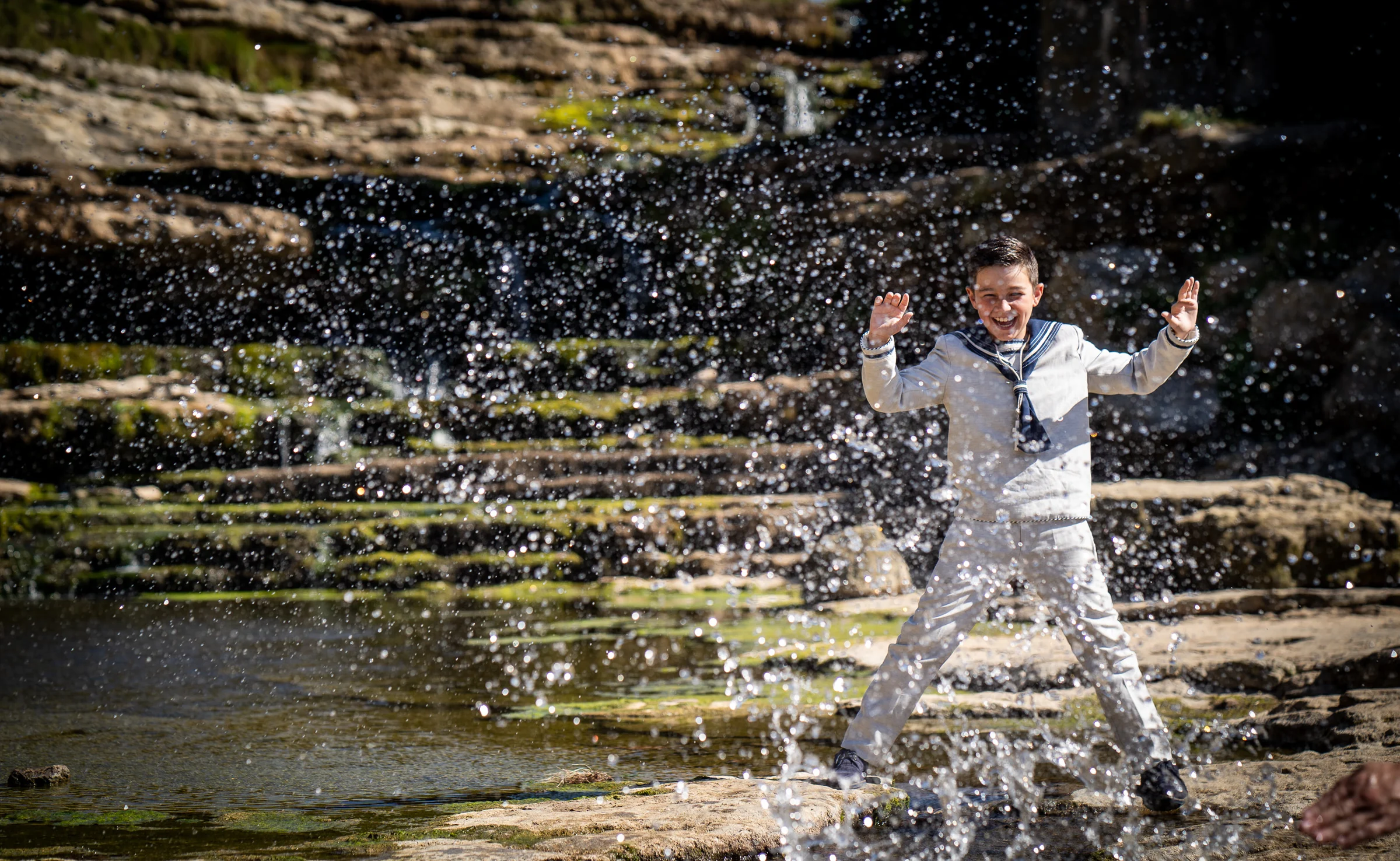 Niño con traje de Primera Comunión juega con el agua frente a una cascada y levanta salpicaduras a su alrededor.