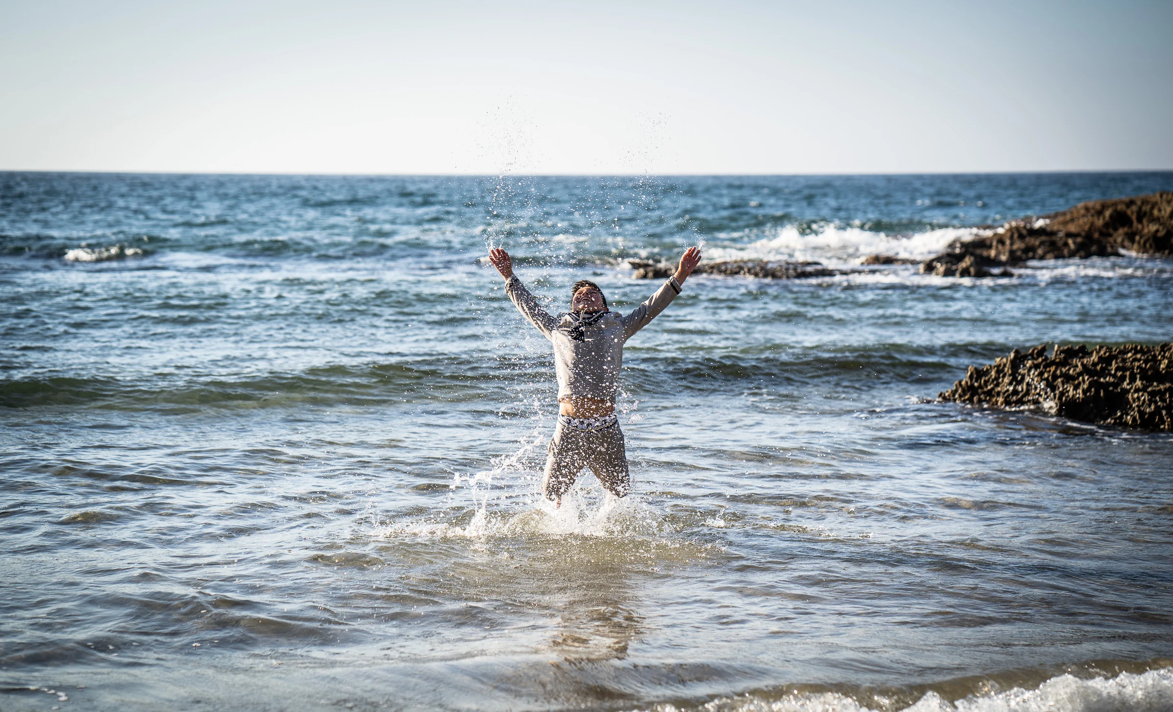 Niño de Primera Comunión entra en el mar con los brazos levantados mientras el agua salpica a su alrededor.