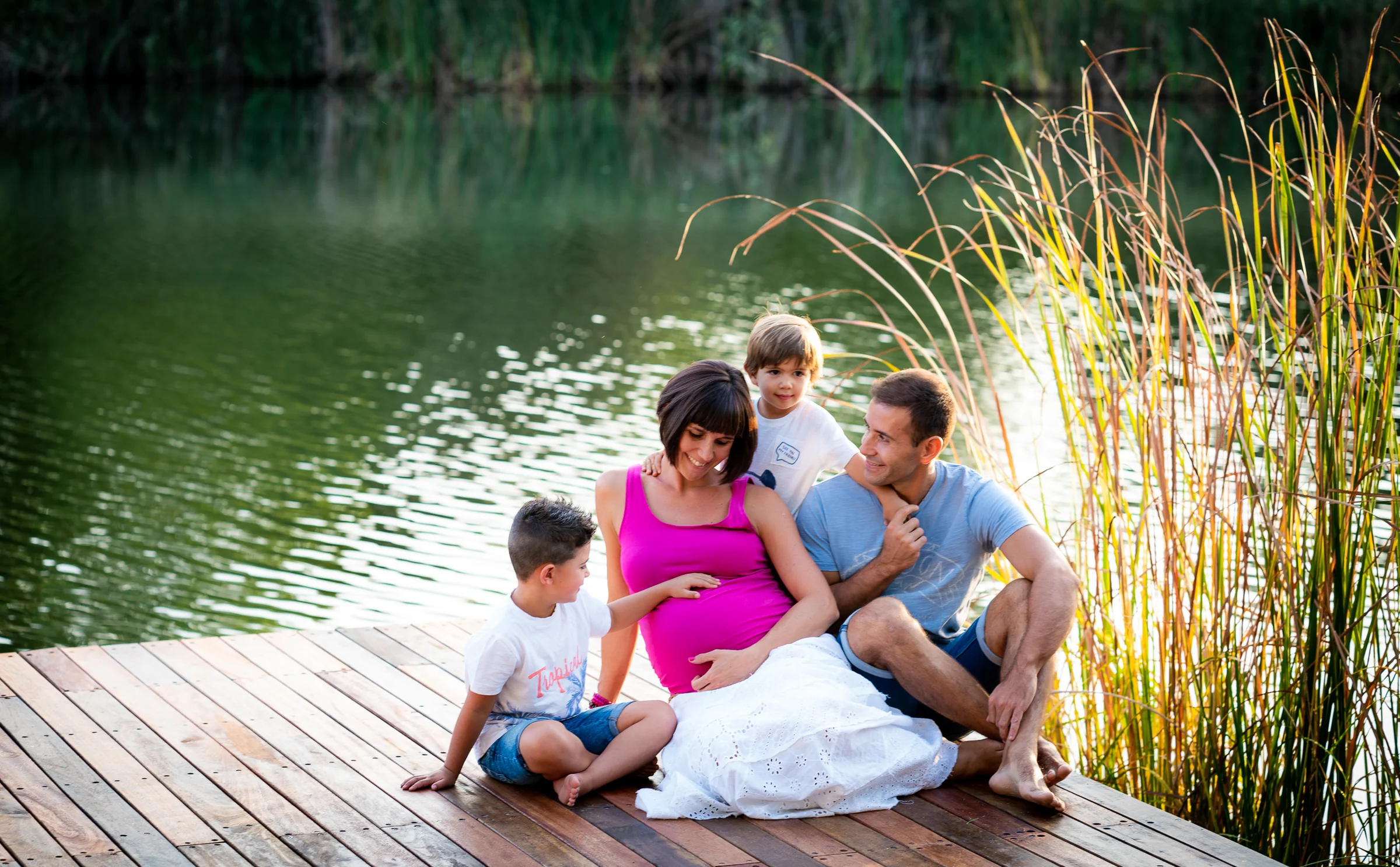 Familia con una mujer embarazada y dos niños sentada en un muelle de madera junto a un lago.