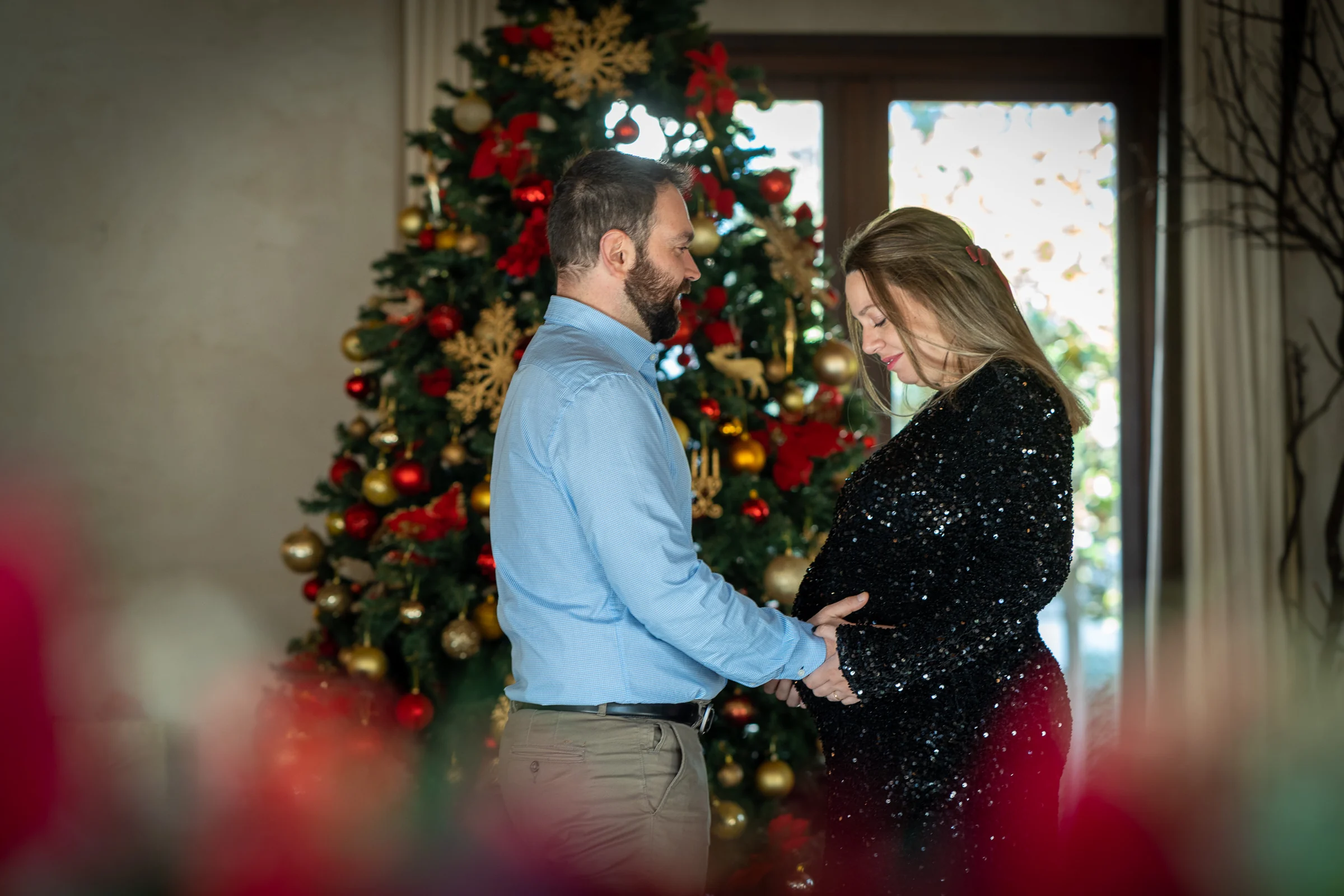 Pareja se mira y acaricia la barriga del embarazo frente a un árbol de Navidad.