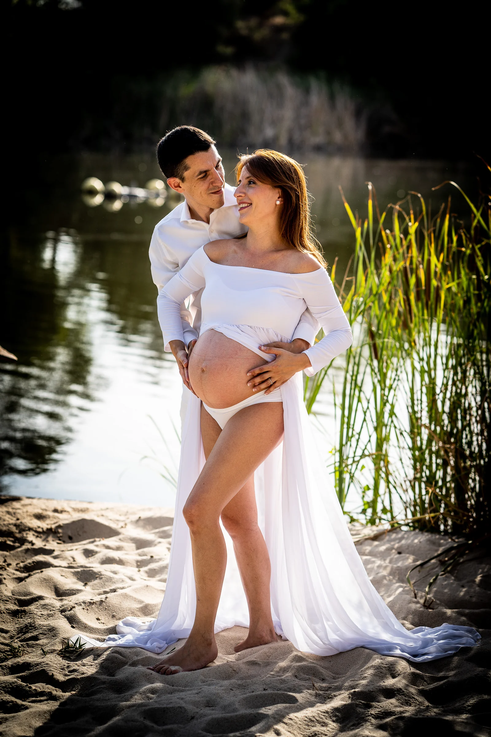 Retrato vertical de una pareja abrazada junto al lago durante una sesión de embarazo con vestido blanco.