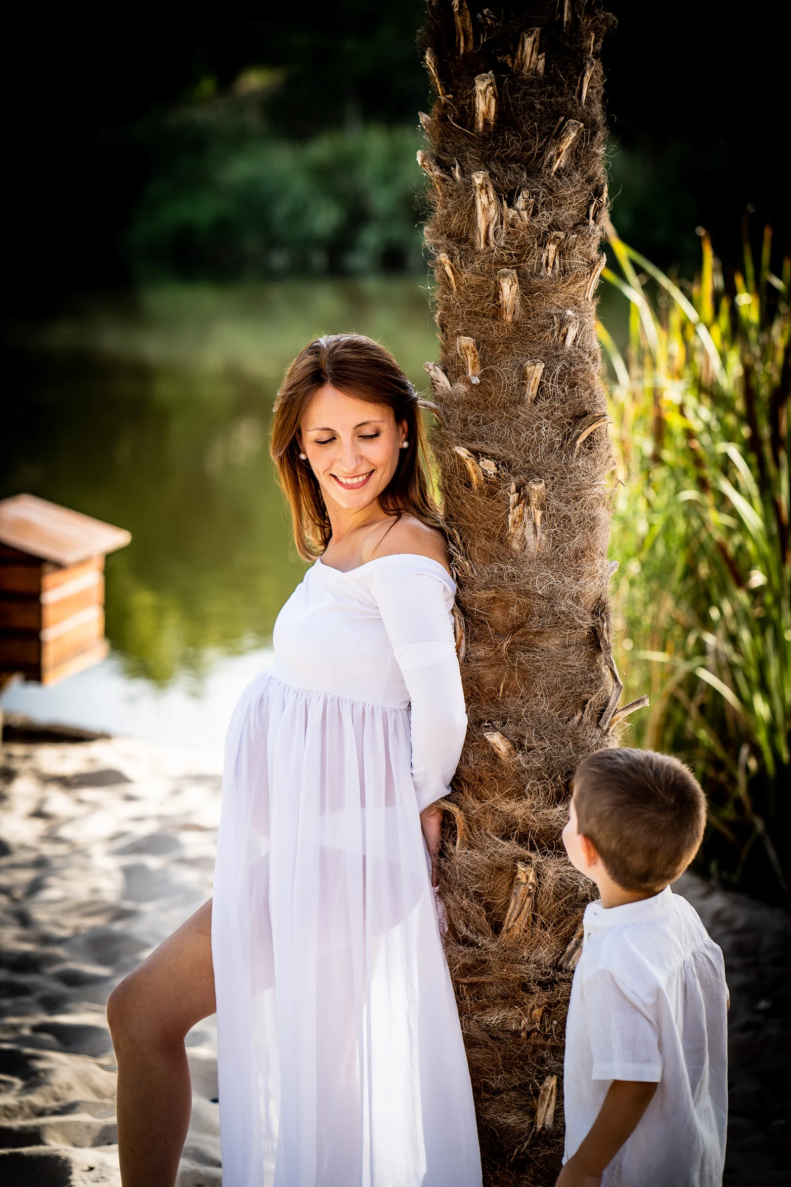 Mujer embarazada con vestido blanco posa junto a una palmera mientras su hijo la mira sonriente.