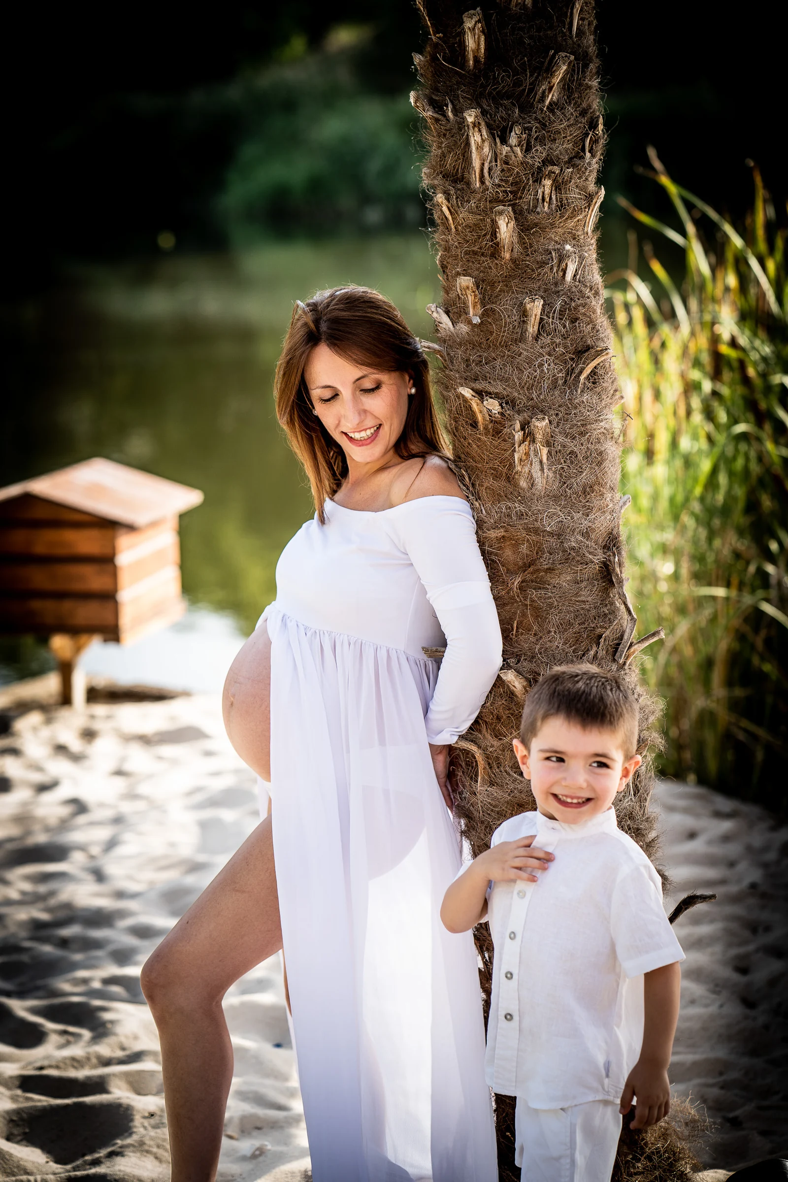 Mujer embarazada con vestido blanco posa junto a una palmera acompañada por su hijo vestido de blanco.