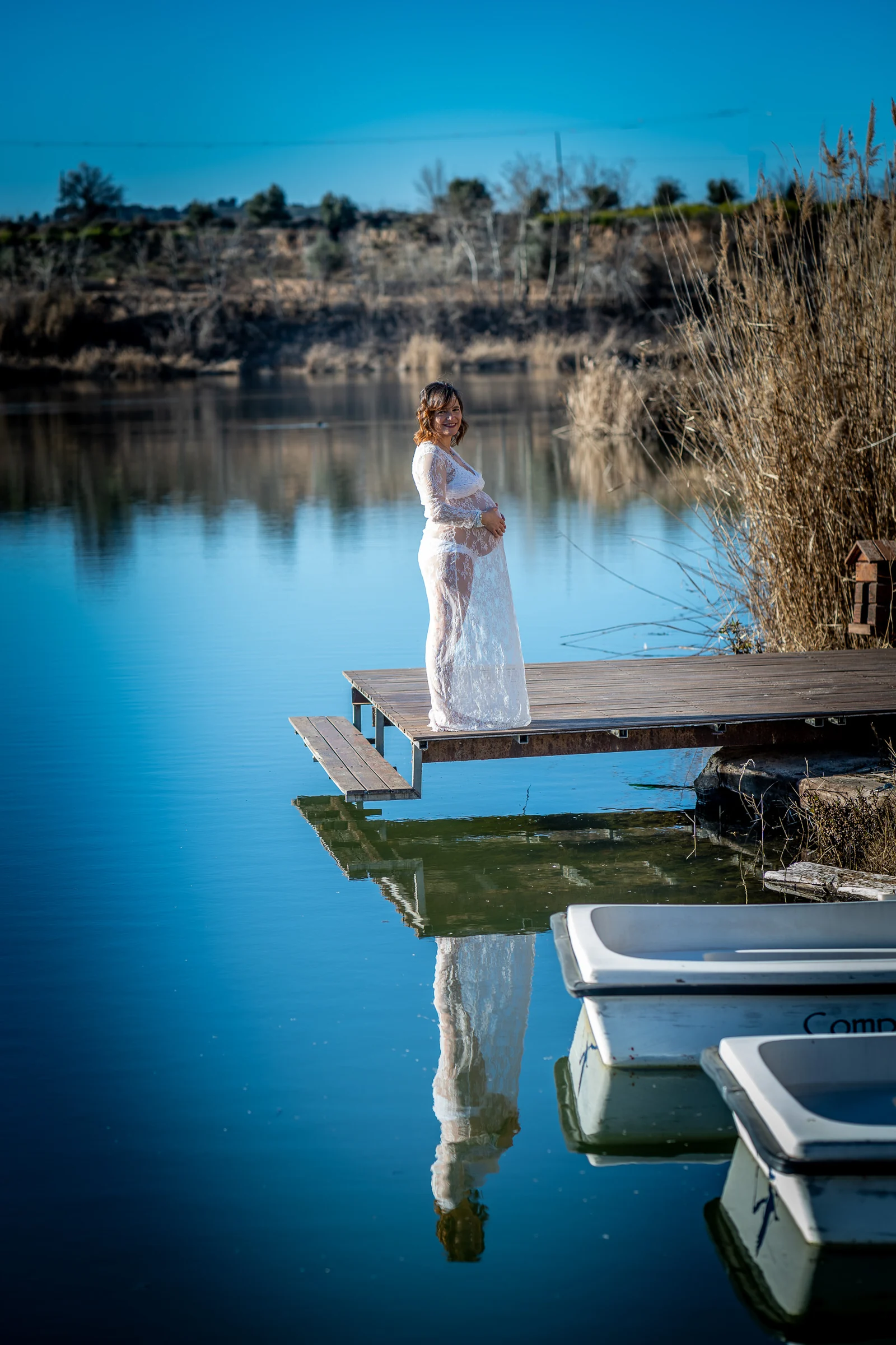 Mujer embarazada con vestido de encaje blanco posa al final de un muelle y se refleja en el agua del lago.