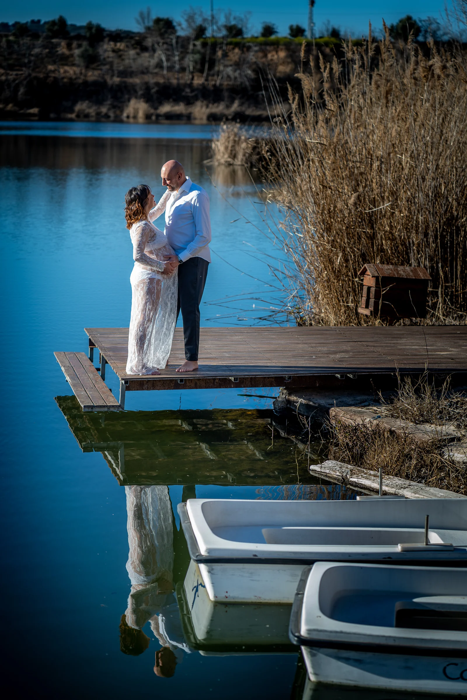 Pareja embarazada posa abrazada en un muelle sobre un lago azul con su reflejo visible en el agua.