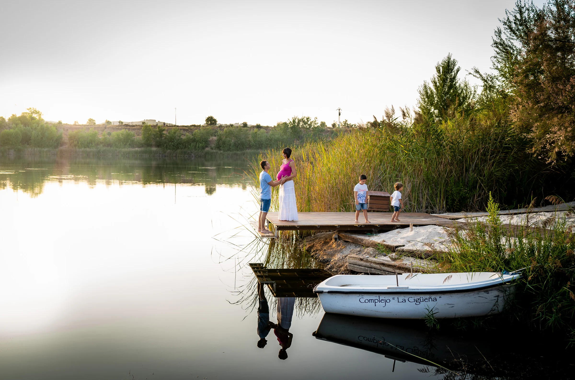 Familia con una mujer embarazada y dos niños posa en un muelle junto a una barca y un lago al atardecer.