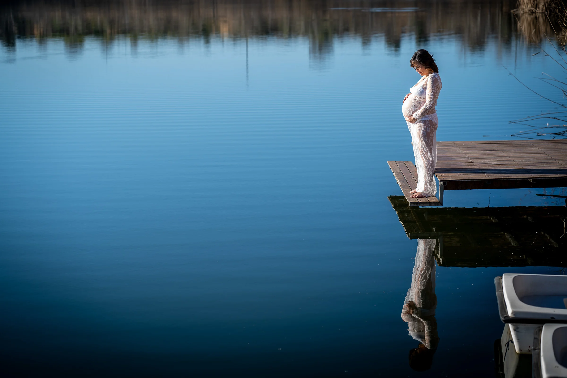 Mujer embarazada con vestido blanco permanece de pie en el extremo de un muelle y se refleja en el agua azul.