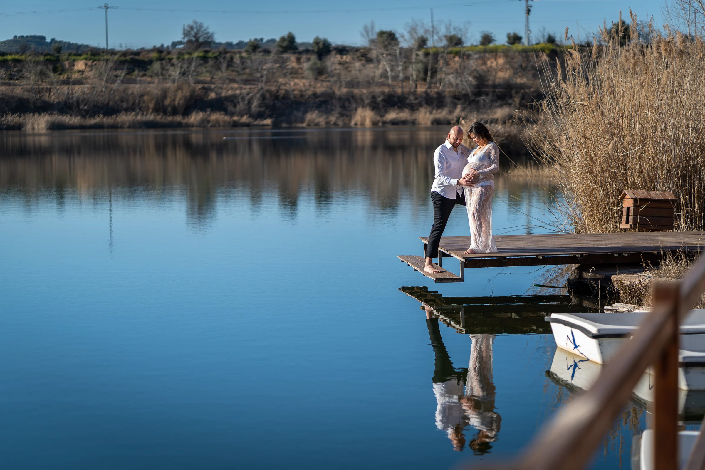 Pareja embarazada se abraza en un muelle junto a un lago tranquilo con su reflejo sobre el agua.