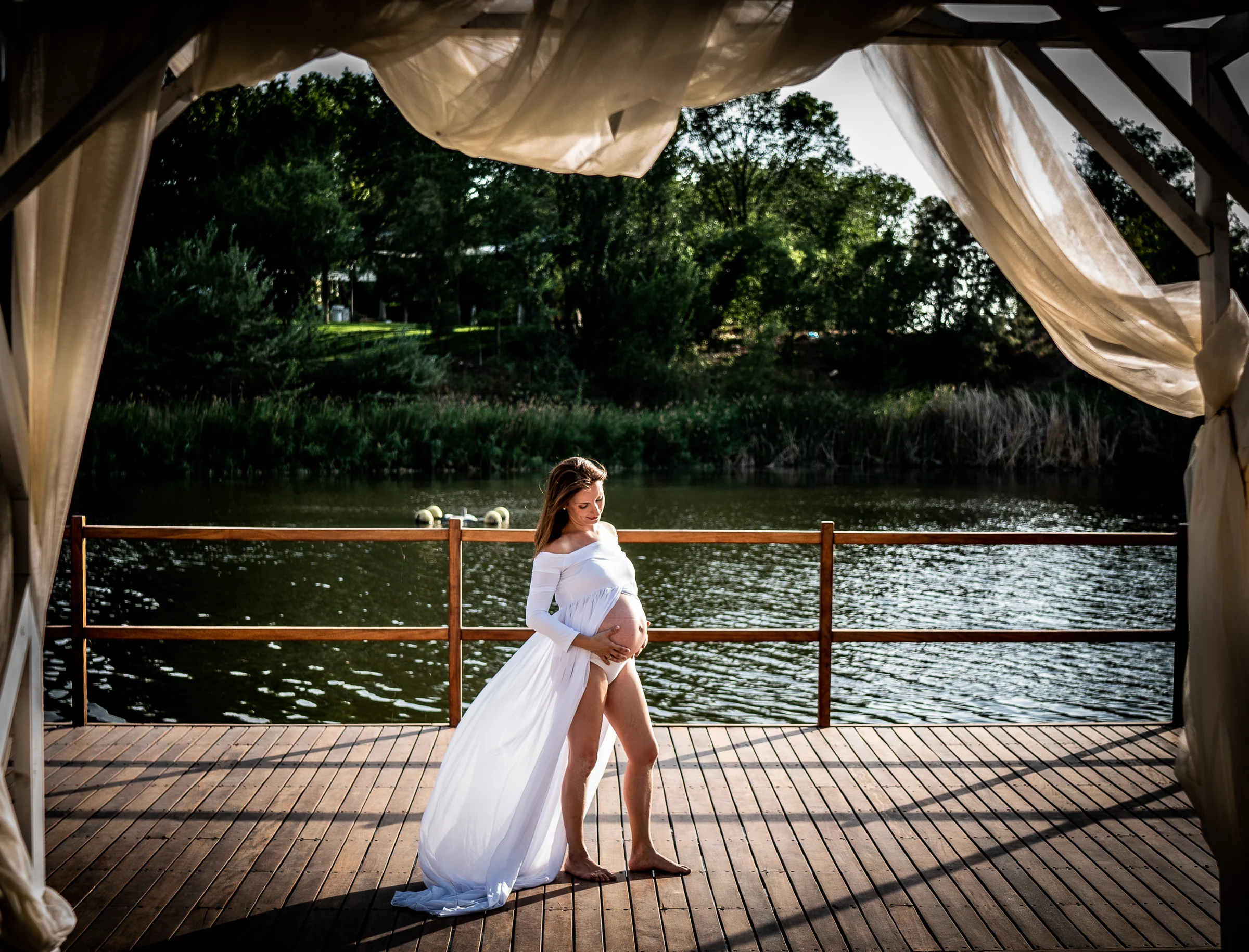 Mujer embarazada con vestido blanco posa bajo una estructura con cortinas frente a un lago.