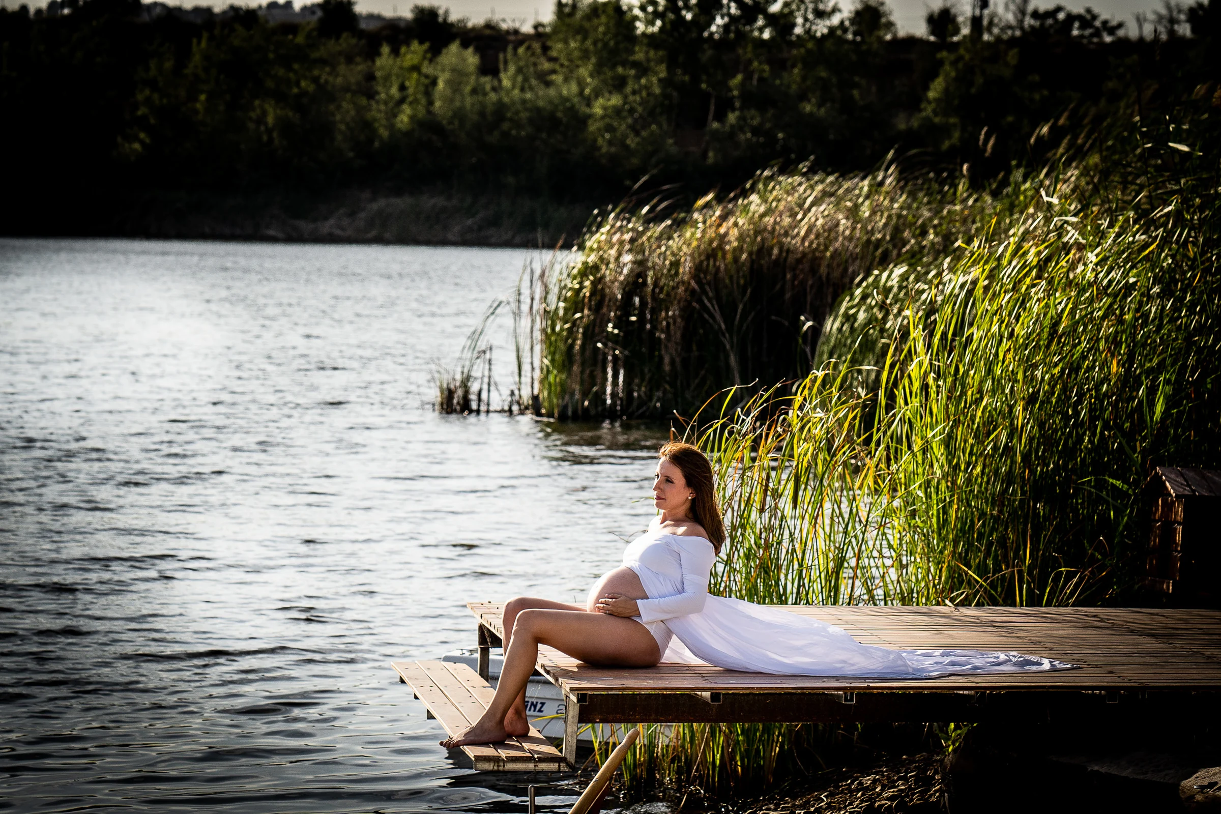 Mujer embarazada con vestido blanco se sienta en el borde de un muelle junto al lago y los carrizos.