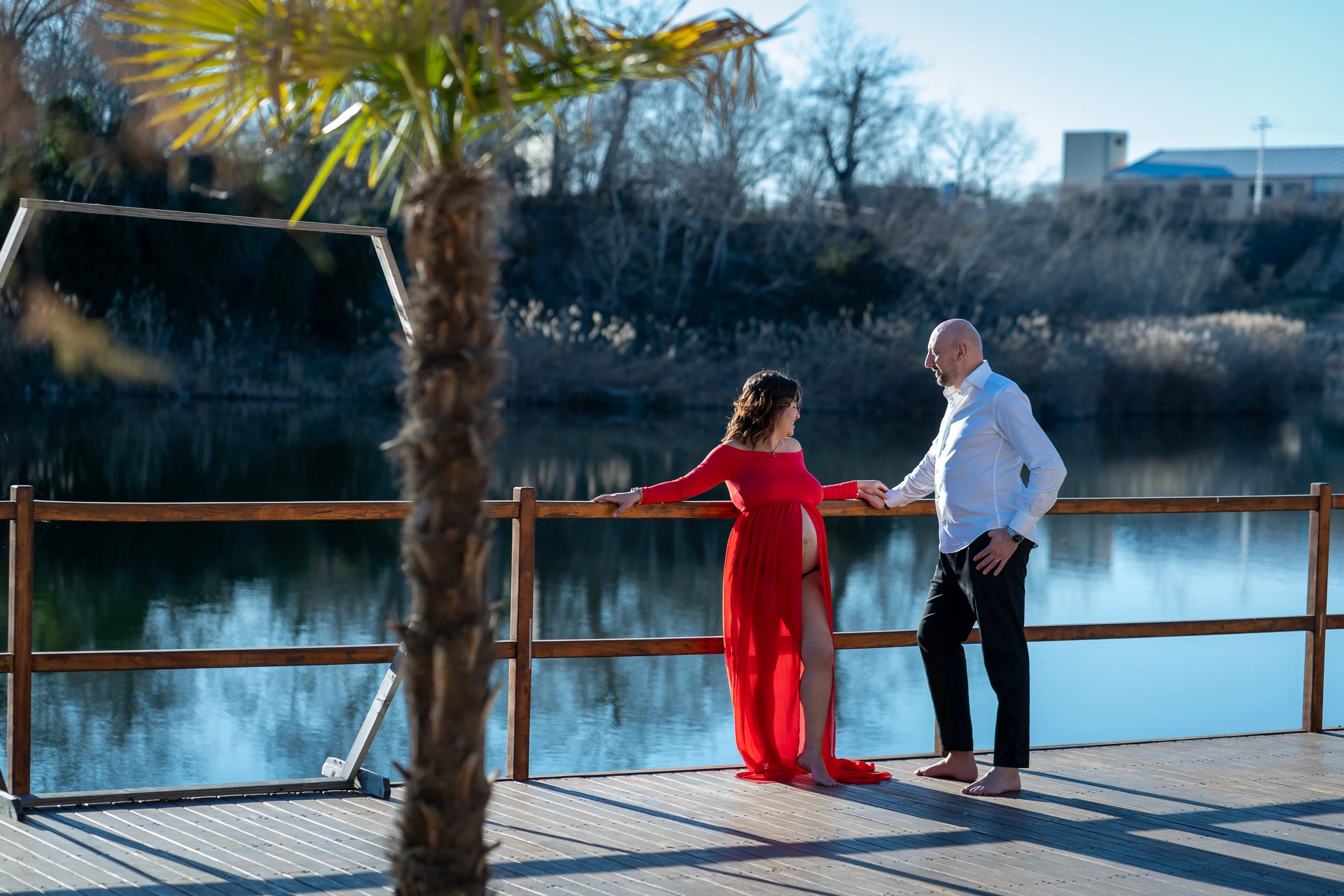 Pareja se toma de la mano junto al lago mientras la mujer embarazada viste un vestido rojo abierto.