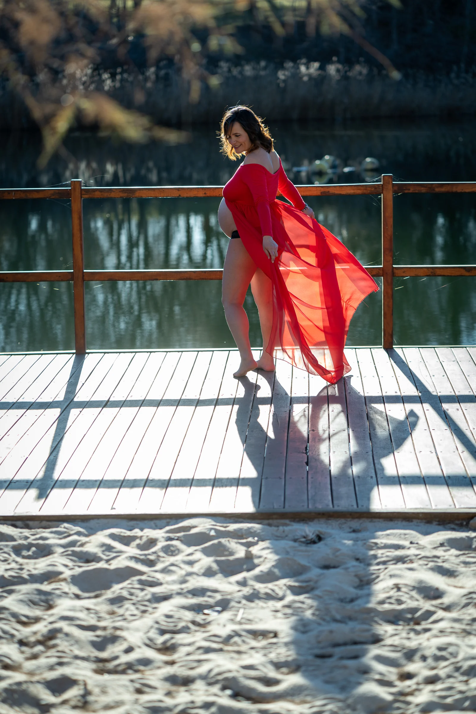 Mujer embarazada con vestido rojo posa de perfil sobre una pasarela de madera y mueve la tela con la mano.