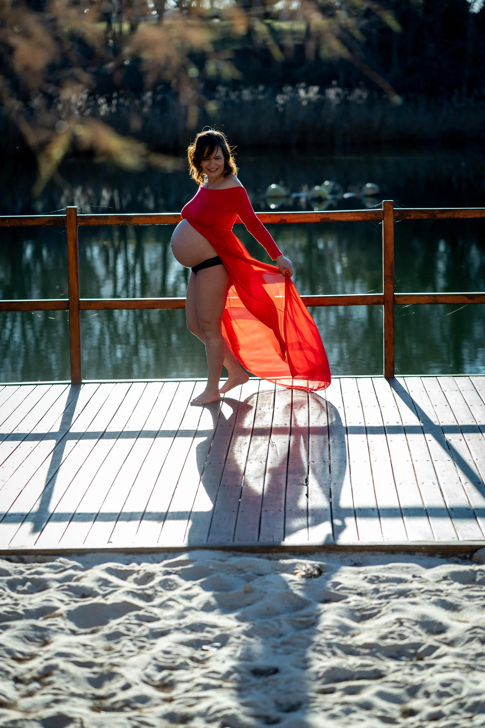 Mujer embarazada con vestido rojo sonríe sobre una pasarela de madera junto al lago.