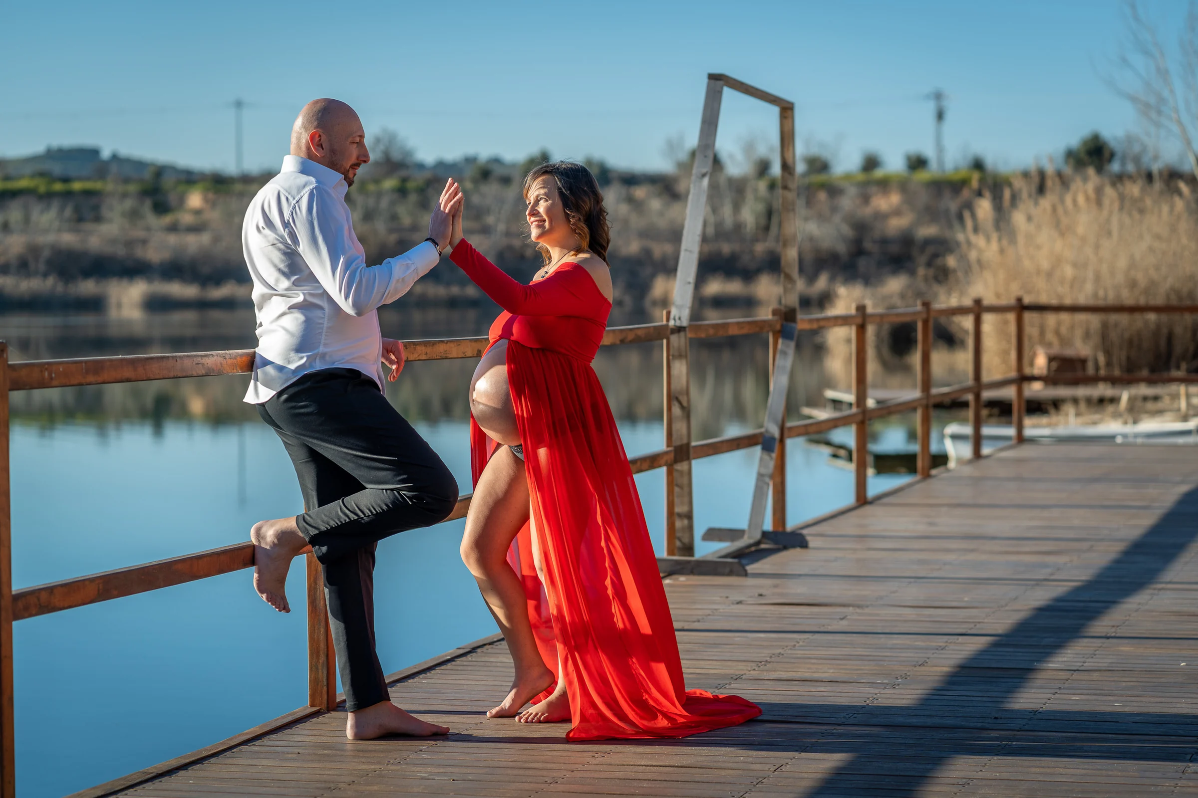 Pareja juega y choca las manos junto al lago durante una sesión de embarazo con vestido rojo.