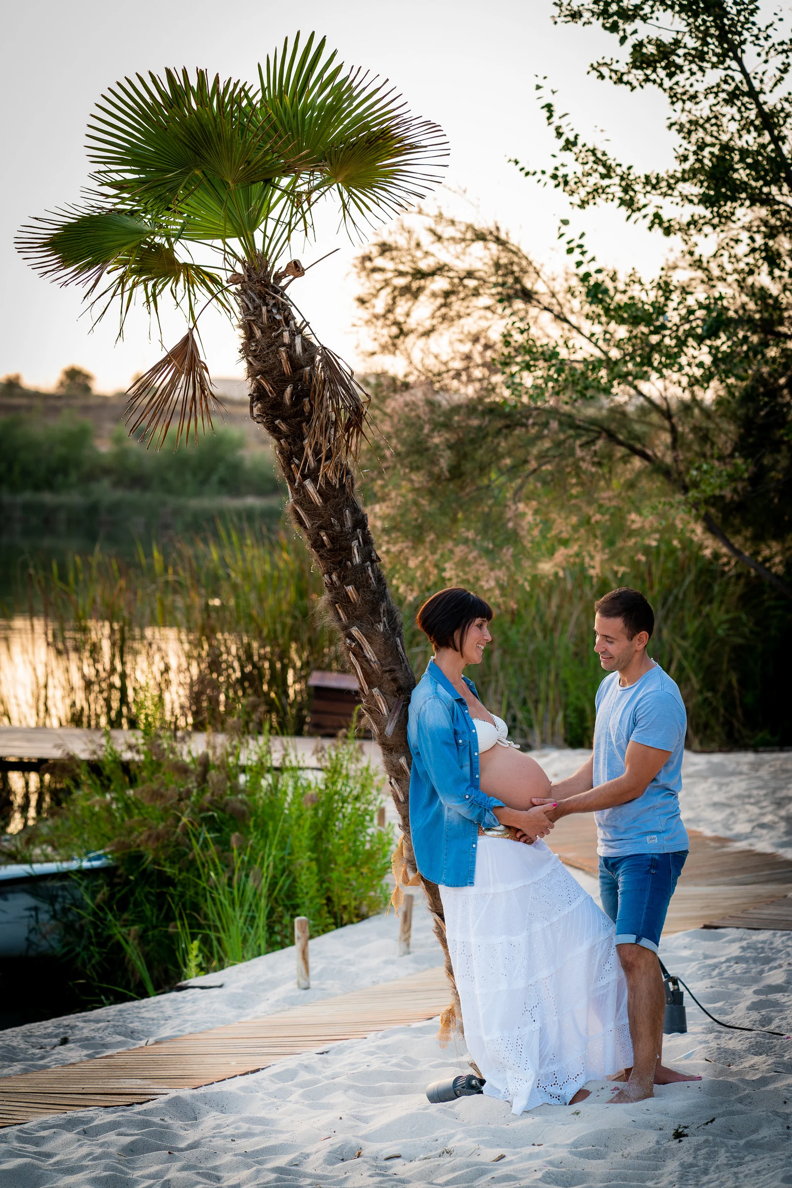 Pareja embarazada posa junto a una palmera en un entorno natural de arena y vegetación.