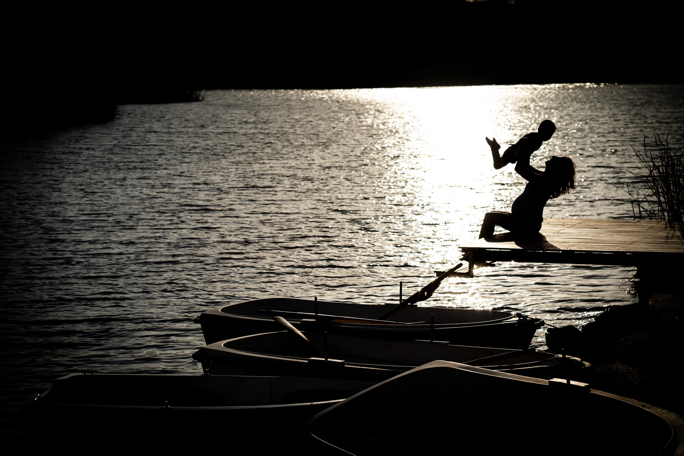 Silueta de una madre embarazada levantando a su hijo en brazos sobre un muelle frente al brillo del lago.