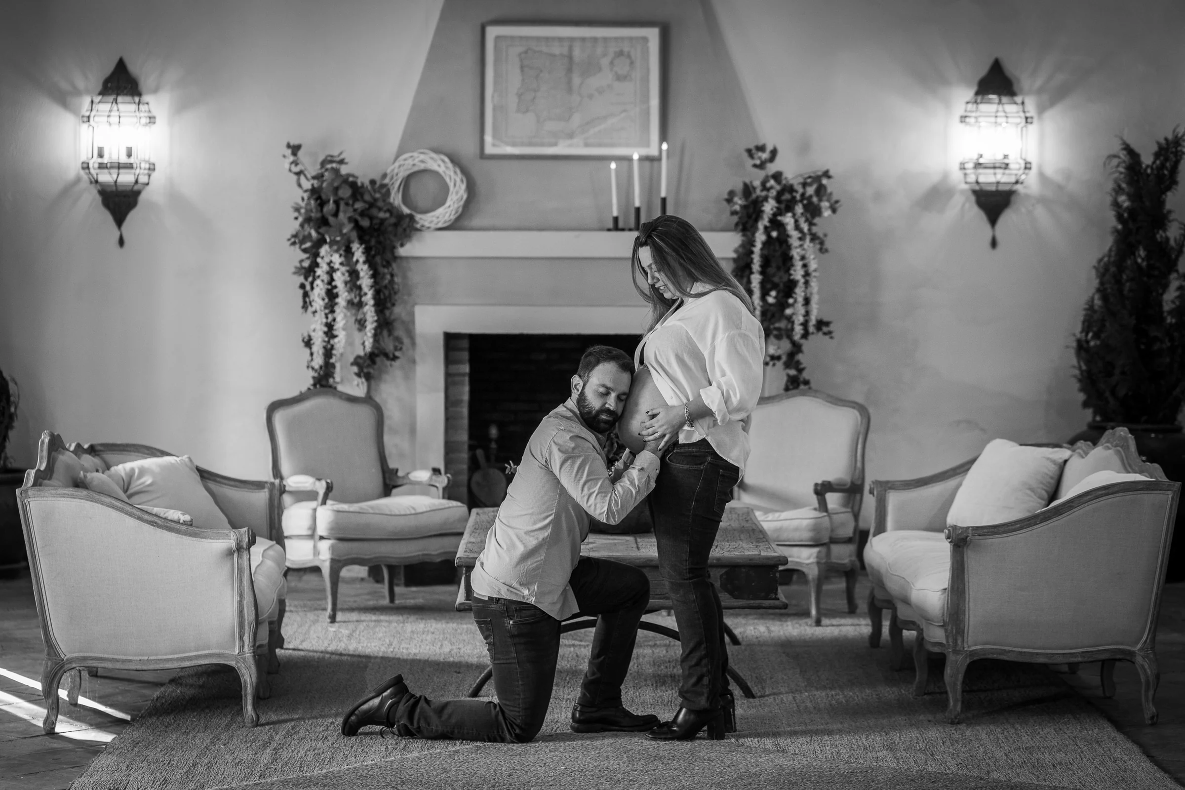 Fotografía en blanco y negro de un hombre arrodillado junto a la barriga de su pareja embarazada en un salón.