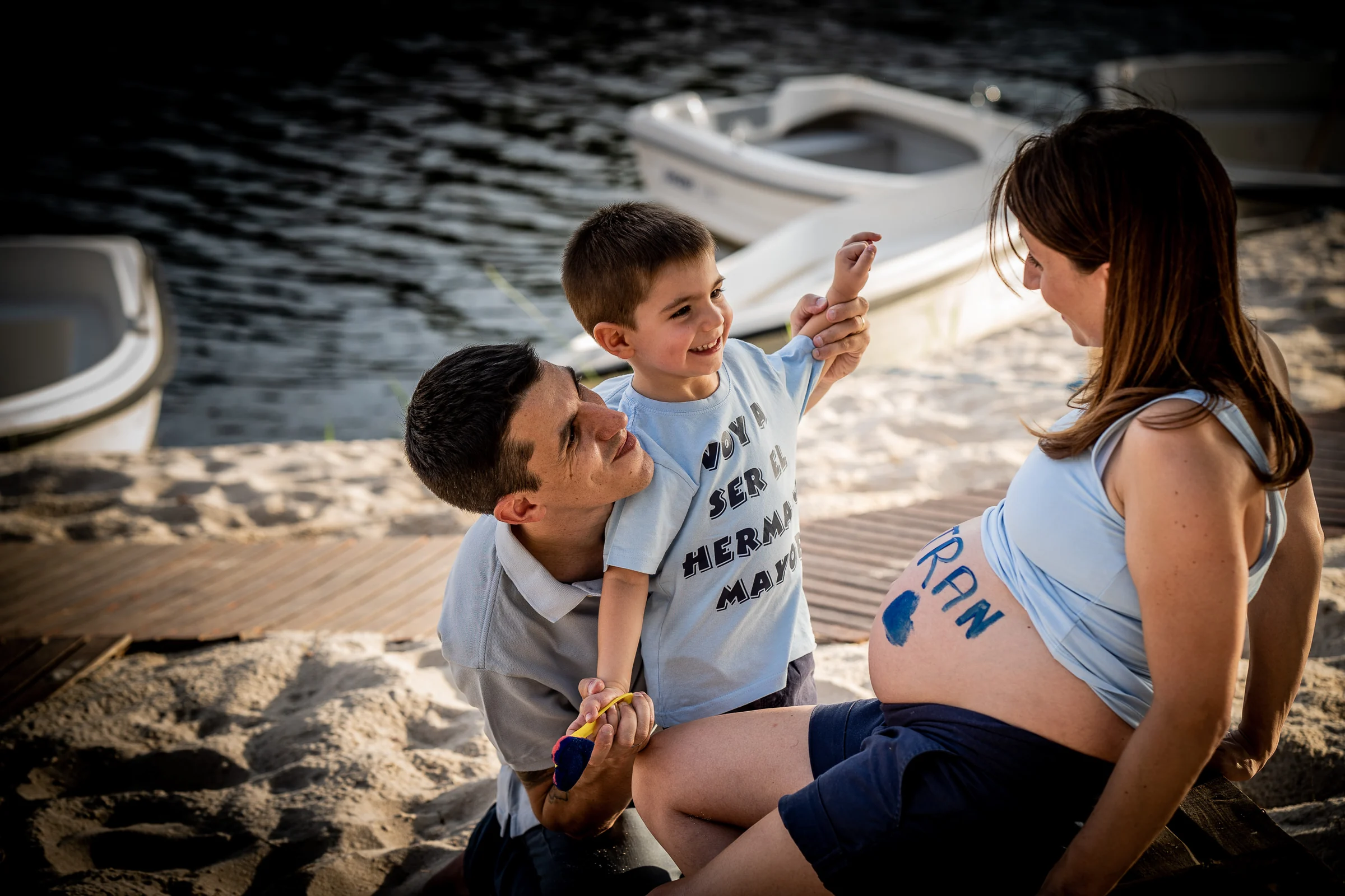Padre y niño juegan junto a una mujer embarazada mientras pintan y escriben sobre su barriga cerca de unas barcas.