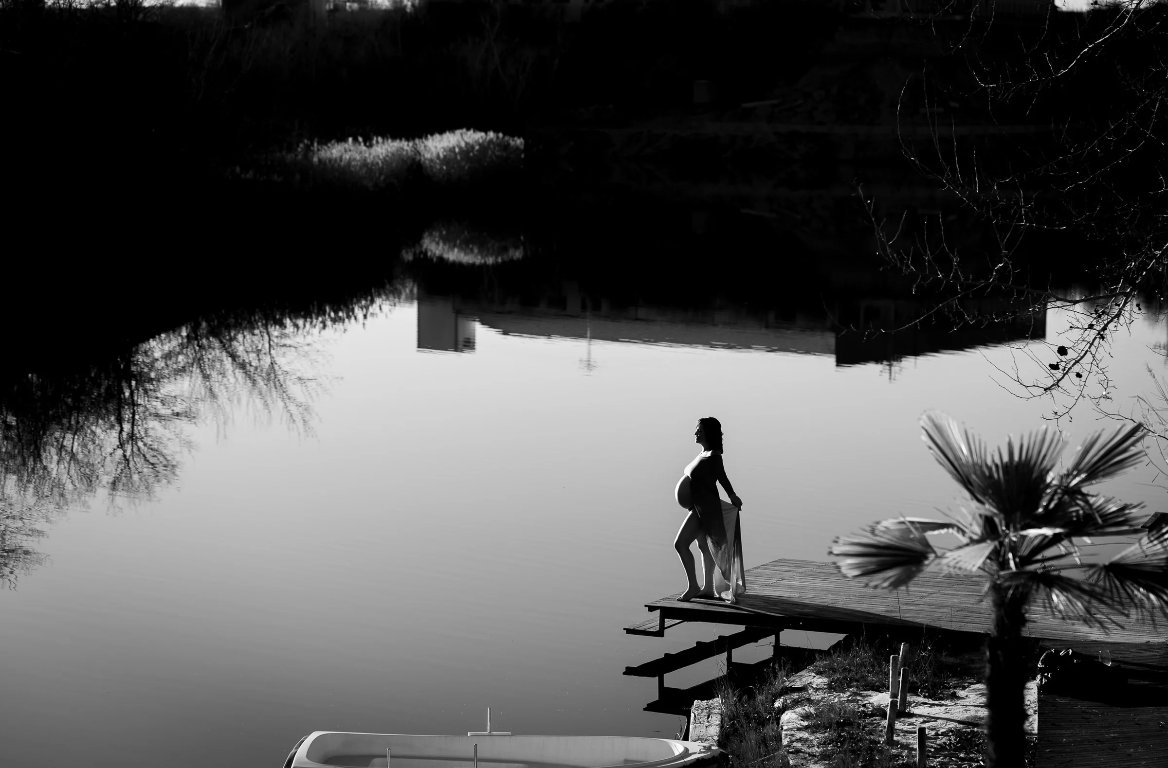Fotografía en blanco y negro de la silueta de una mujer embarazada sobre un muelle junto a un lago en calma.