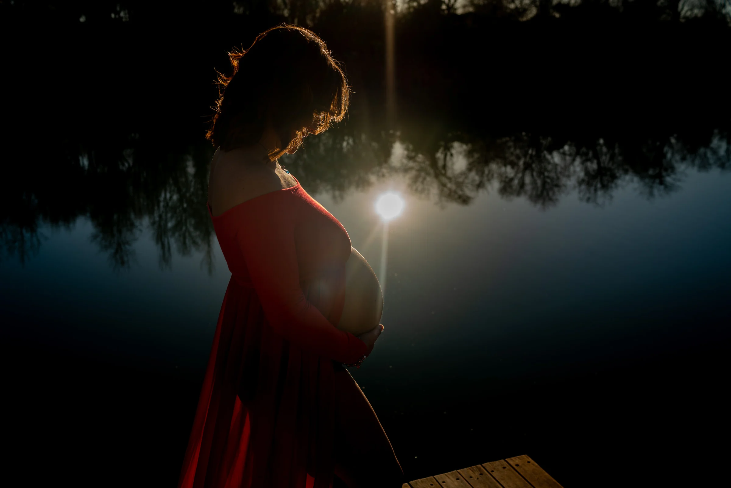 Silueta parcial de una mujer embarazada con vestido rojo frente al reflejo del sol sobre el lago.