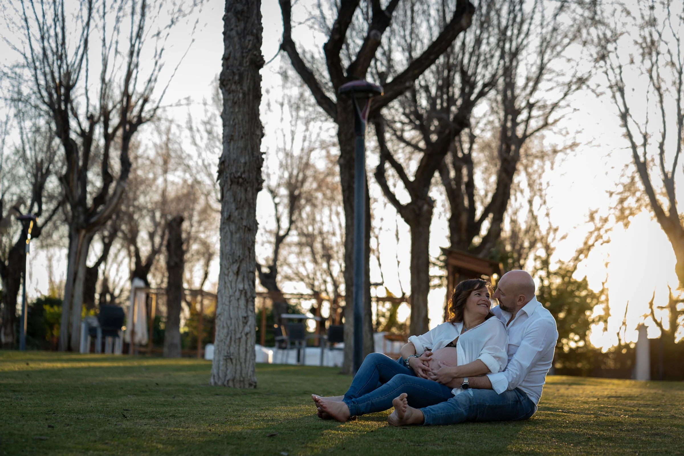 Pareja embarazada sentada sobre el césped se abraza y mira al atardecer entre árboles.